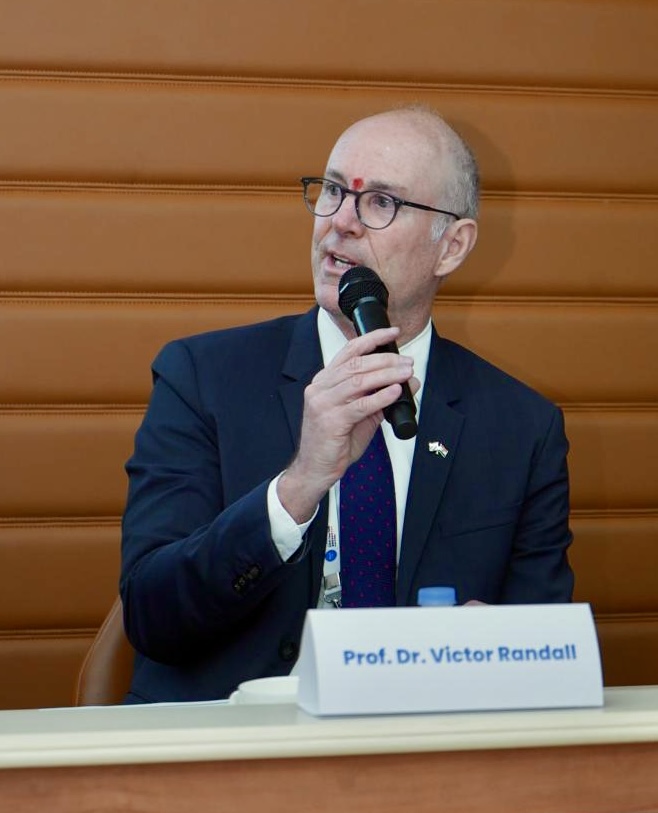 Ein Mann im Anzug und mit Brille, mit einem roten Fleck auf der Stirn, spricht im Sitzen in ein Mikrofon. Auf einem Namensschild vor ihm steht "Prof. Dr. Victor Randall", ein Experte für Finanzmanagement. Im Hintergrund ist eine braune, gepolsterte Verkleidung zu sehen. at Hochschule Coburg