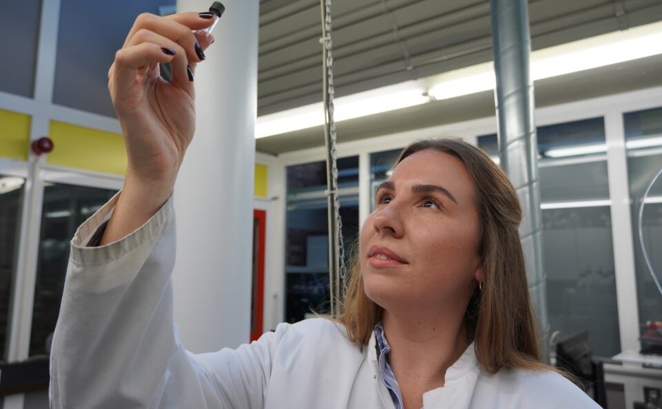 Eine Frau in einem weißen Laborkittel untersucht in einem Labor der Hochschule ein kleines Fläschchen, dessen Inhalt sie im Rahmen der Forschung zur Neuen Therapie bei hellem Licht genau unter die Lupe nimmt. at Hochschule Coburg