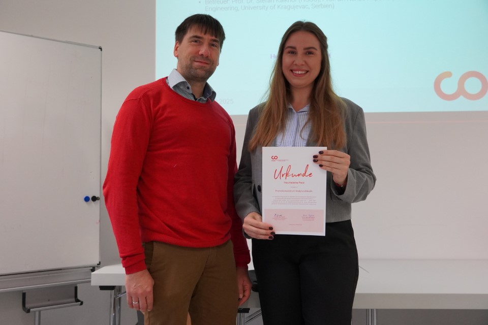 Ein Mann in einem roten Pullover steht neben einer lächelnden Frau in einem Blazer, die ein Zertifikat für eine neue Therapie gegen Krebs in der Hand hält, in einem hellen Raum der Hochschule mit einer Tafel und einem Projektor im Hintergrund. at Hochschule Coburg