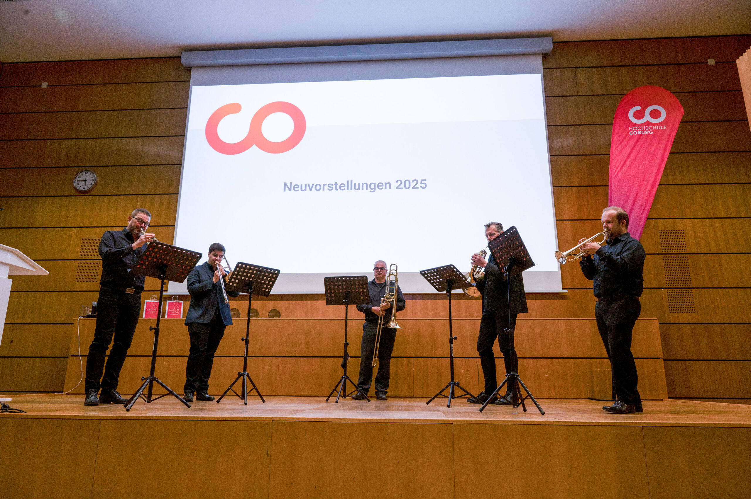 Fünf Musiker in schwarzen Anzügen spielen mit Blechblasinstrumenten auf der Bühne der Hochschule. Hinter ihnen zeigt eine große Leinwand "Neuvorstellungen 2025" und ein rotes Logo in der holzgetäfelten Aula des Dies academicus. at Hochschule Coburg