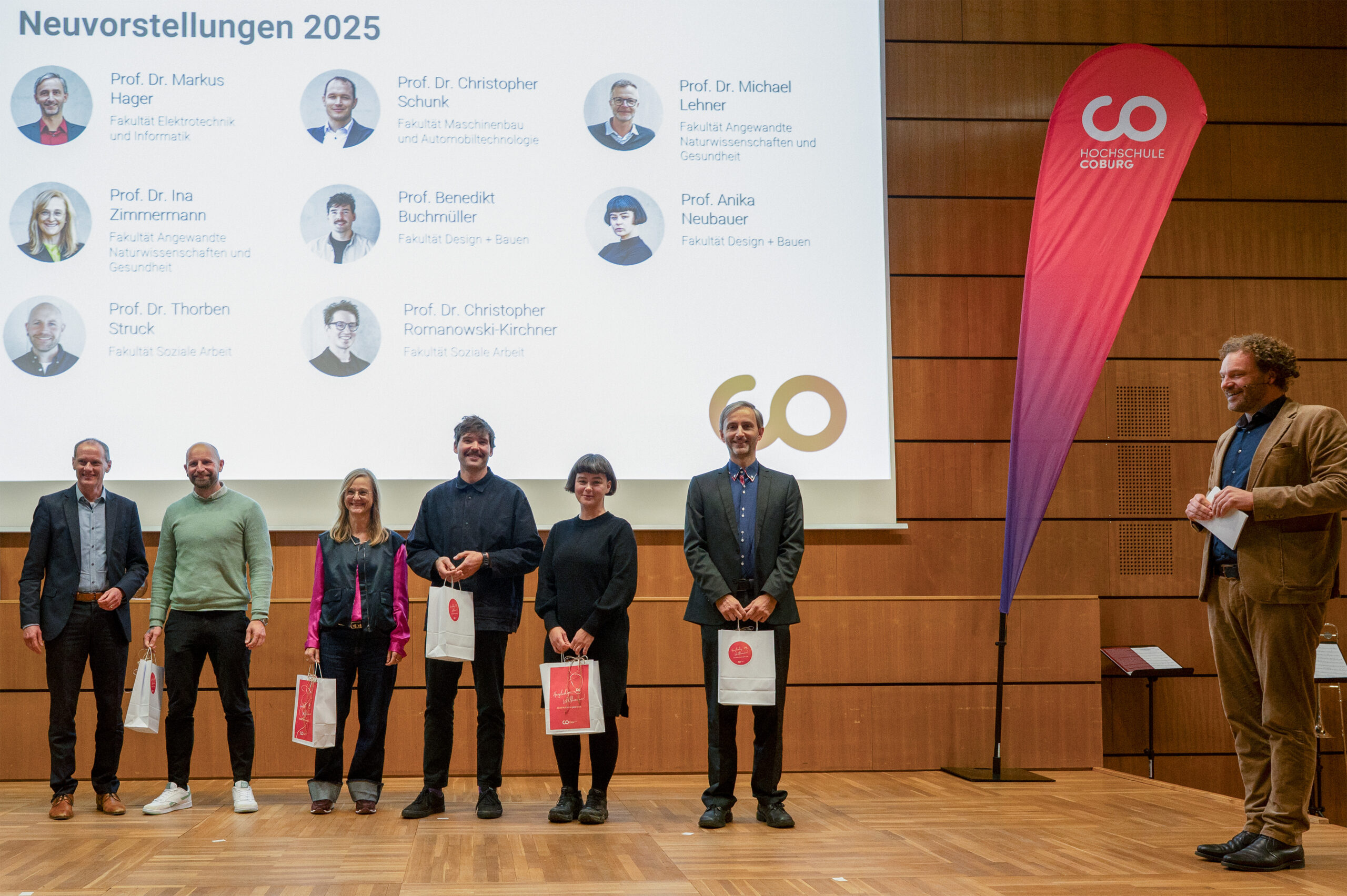 Sieben Personen stehen auf einer Holzbühne und halten Geschenktüten in der Hand, während ein Mann zu ihnen spricht. Hinter ihnen zeigt ein Bildschirm die Namen und Fotos der Dozenten sowie ein rotes Banner der Hochschule Coburg - eine akademische Szene, die wahrscheinlich vom Dies academicus 2025 stammt. at Hochschule Coburg