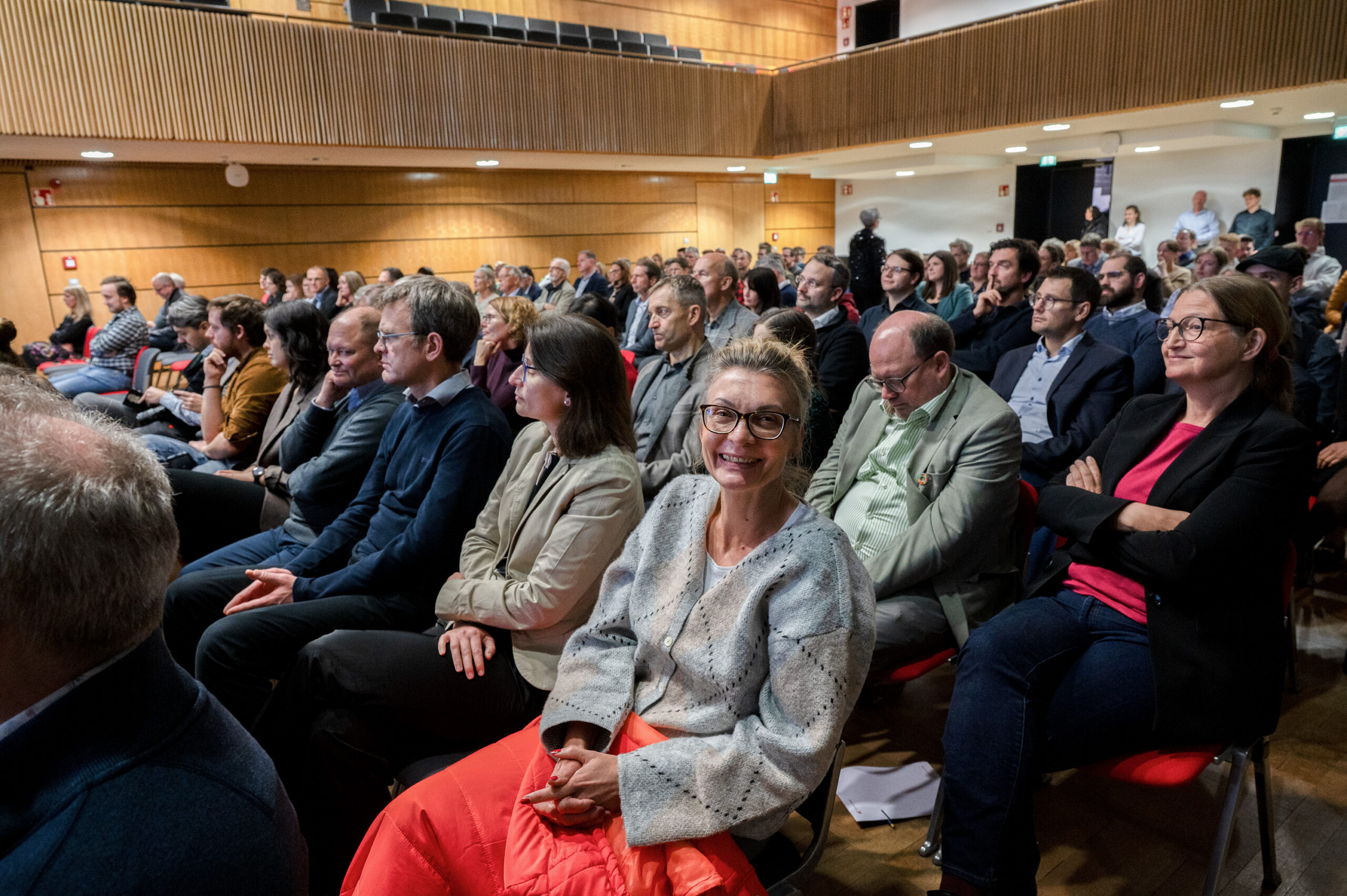 Eine Frau im hellen Pullover und mit Brille lächelt in die Kamera, während sie während des Dies academicus 2025 inmitten eines großen Publikums in der modernen Aula der Hochschule sitzt. Die meisten Menschen konzentrieren sich auf ein Ereignis, das außerhalb des Blickfelds stattfindet. at Hochschule Coburg