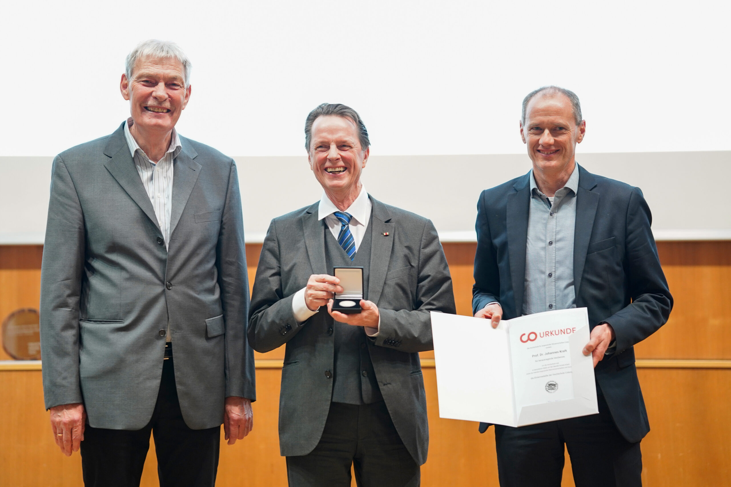 Drei Männer in Anzügen stehen lächelnd in einem Innenraum bei einer Preisverleihung an einer Hochschule - Teil des Dies academicus 2025. Der Mann in der Mitte hält eine Medaille in einer Schachtel, während der Mann rechts eine Urkunde zeigt. at Hochschule Coburg