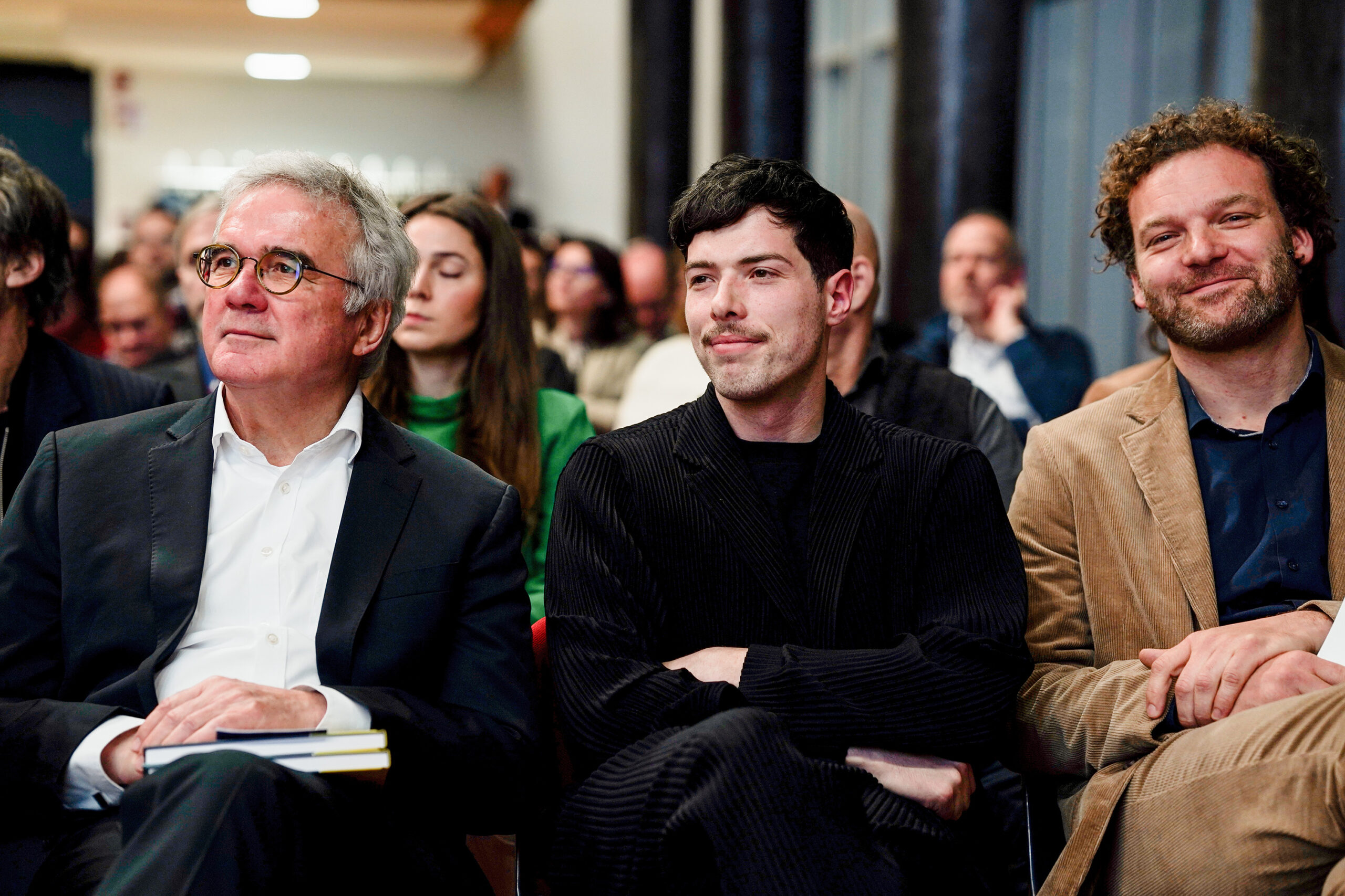 Drei Männer sitzen nebeneinander in einer Audienz an der Hochschule Dies academicus 2025, gekleidet in Anzug und Jackett, und wirken aufmerksam und nachdenklich. Im Hintergrund sind weitere Personen zu sehen, die sich unscharf abzeichnen. at Hochschule Coburg