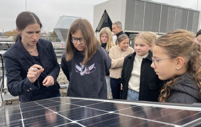 Eine Frau erklärt einer Gruppe aufmerksamer Schüler, wie ein Solarmodul funktioniert. Sie inspiriert jedes Mädchen auf dem Dach, die Zukunft der Energie zu entdecken, während im Hintergrund große Industrieanlagen zu sehen sind. at Hochschule Coburg