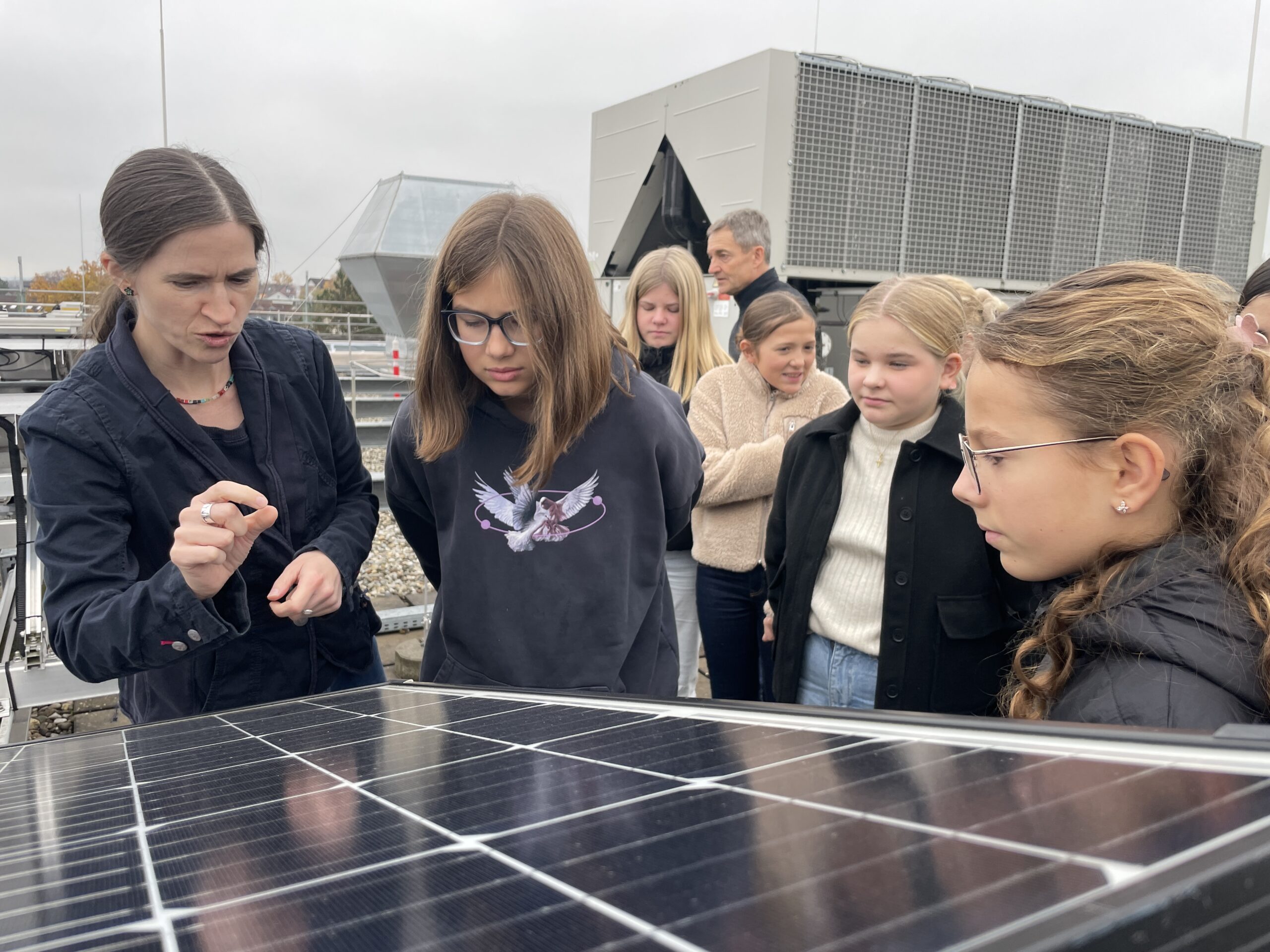 Eine Frau erklärt einer Gruppe aufmerksamer Schüler, wie ein Solarmodul funktioniert. Sie inspiriert jedes Mädchen auf dem Dach, die Zukunft der Energie zu entdecken, während im Hintergrund große Industrieanlagen zu sehen sind. at Hochschule Coburg