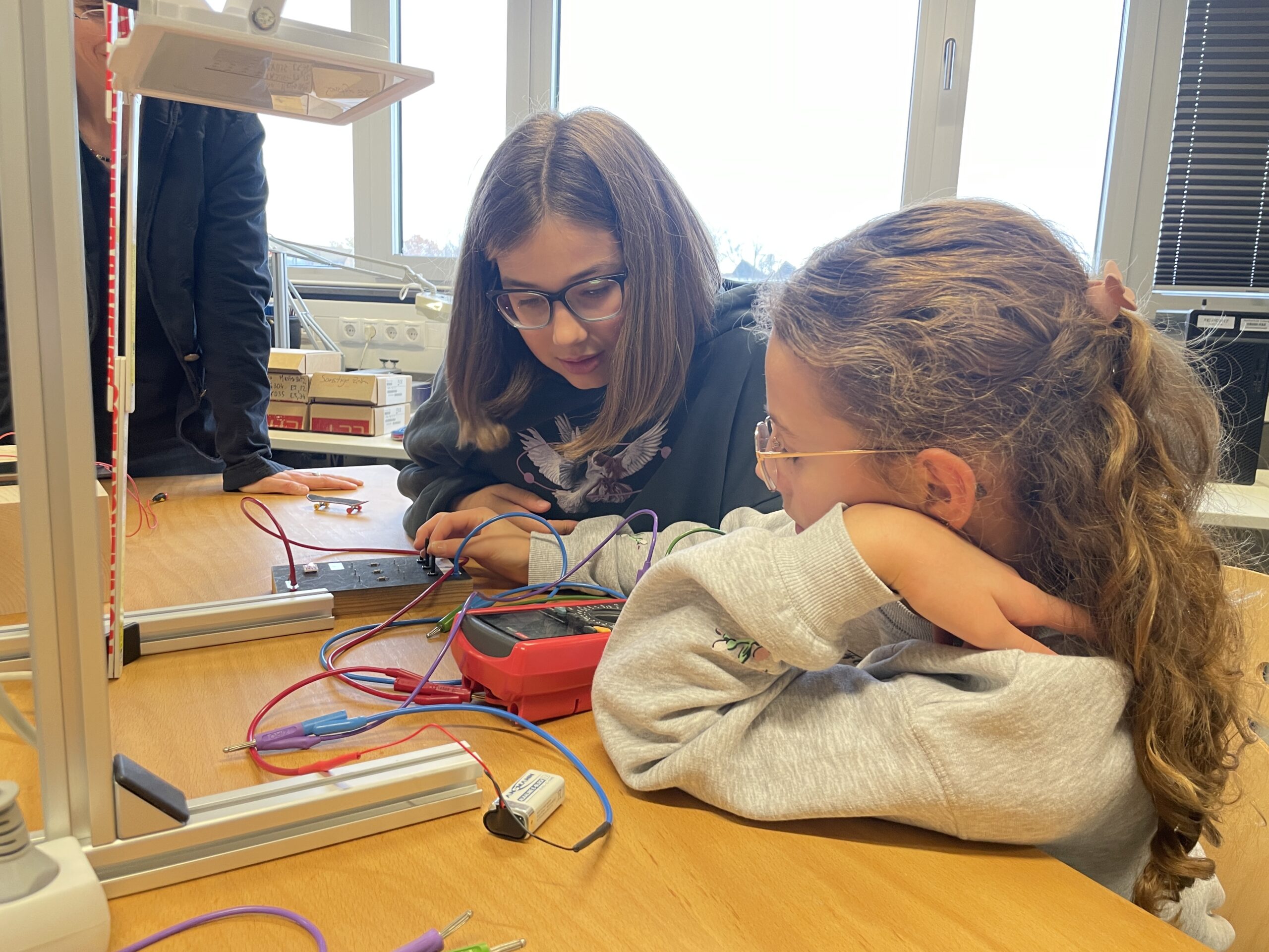 Zwei Mädchen sitzen an einem Holztisch und arbeiten konzentriert an einem Elektronikprojekt mit Drähten und einem Multimeter, vereint in ihrer Mission, die Zukunft zu gestalten, in einem hellen Klassenzimmer mit Fenstern im Hintergrund. at Hochschule Coburg