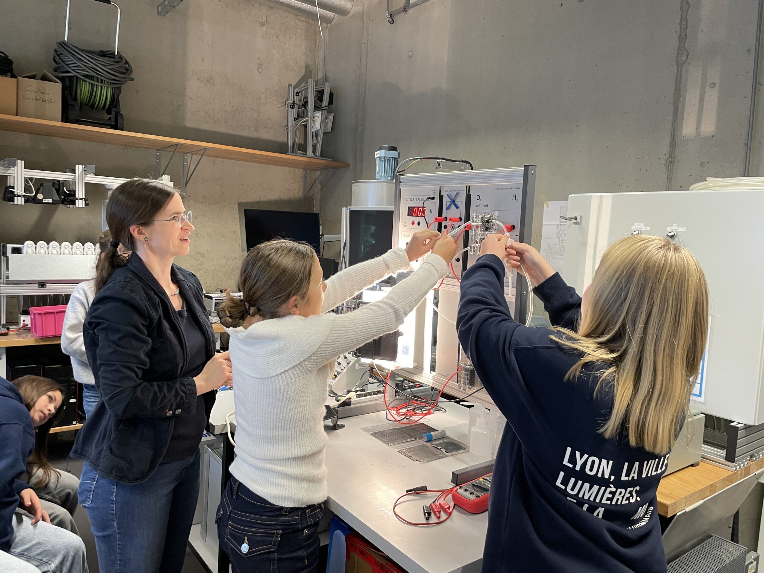 Drei Frauen arbeiten gemeinsam an einem Elektronikprojekt in einem Labor und schließen Drähte an eine Schalttafel an. Die Frau auf der linken Seite beobachtet und lächelt, während die anderen sich auf ihre Aufgabe konzentrieren. Im Hintergrund sitzen ein paar Leute. at Hochschule Coburg