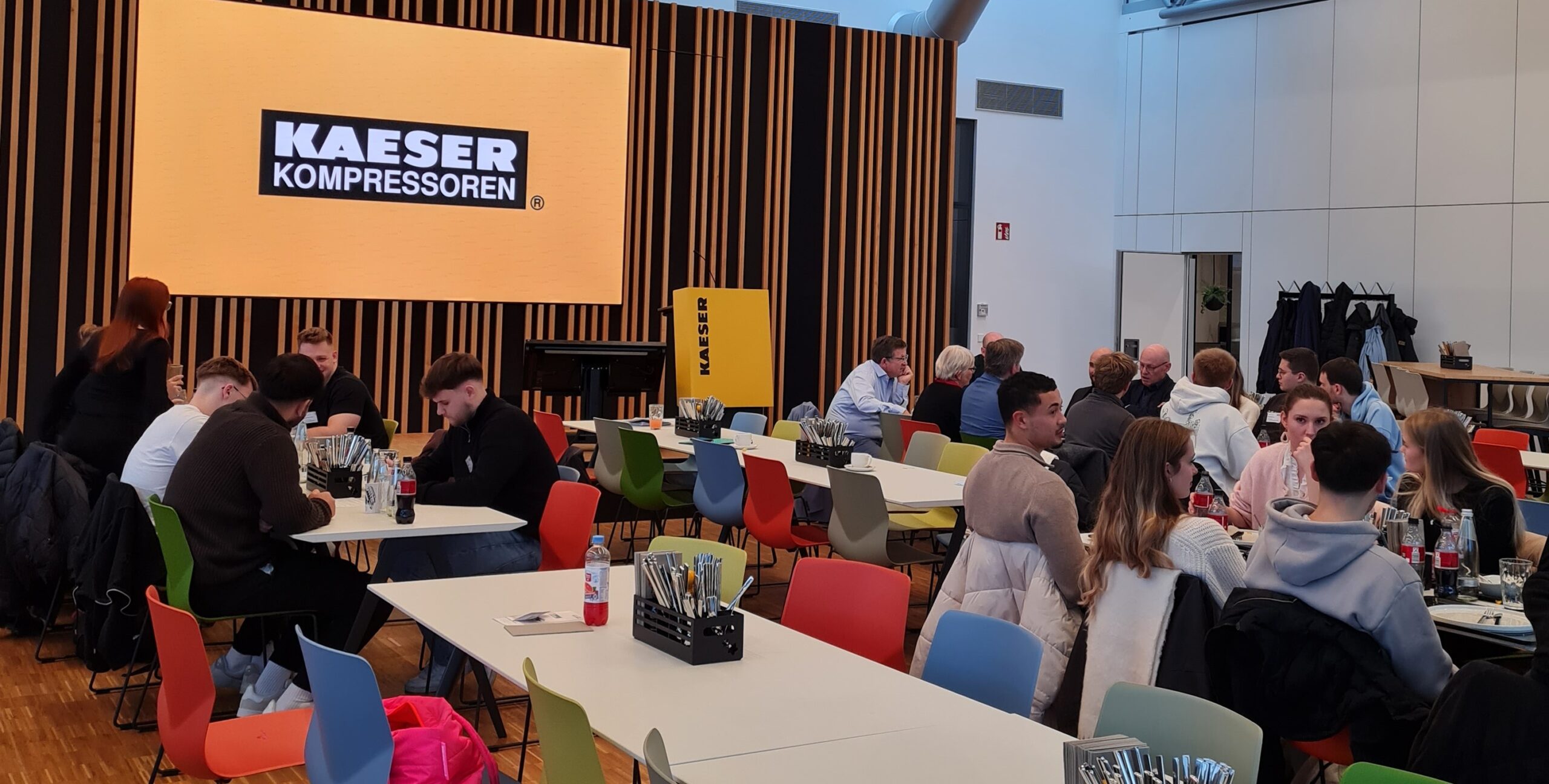 In einer modernen Cafeteria mit bunten Stühlen sitzen Menschen an Tischen, diskutieren über Wirtschaft und essen. Auf einem großen Bildschirm ist das Logo "KAESER KOMPRESSOREN" in Weiß auf schwarz-gelbem Hintergrund zu sehen. at Hochschule Coburg