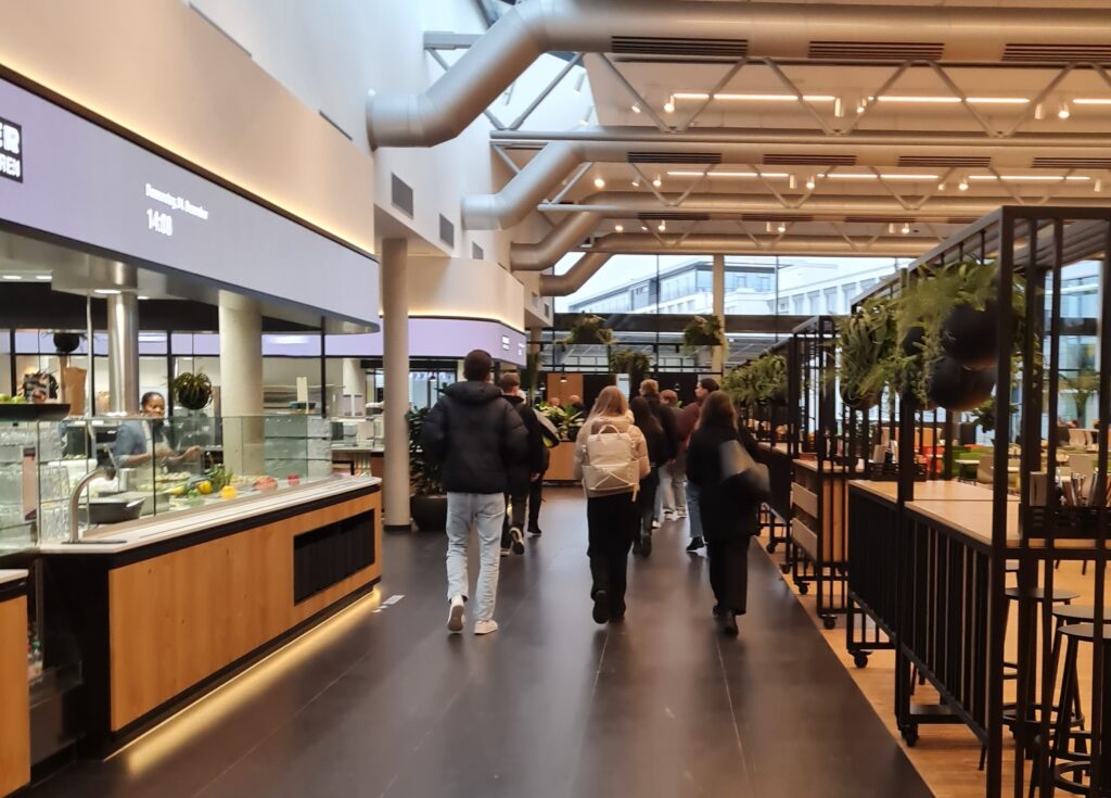Eine Gruppe von Menschen geht durch eine moderne, geräumige Cafeteria mit hohen Decken, großen Fenstern, Pflanzen und einer Essensausgabe auf der linken Seite. Die Atmosphäre ist hell und zeitgemäß - ideal für Gespräche über Wirtschaft oder Versicherungsmanagement. at Hochschule Coburg
