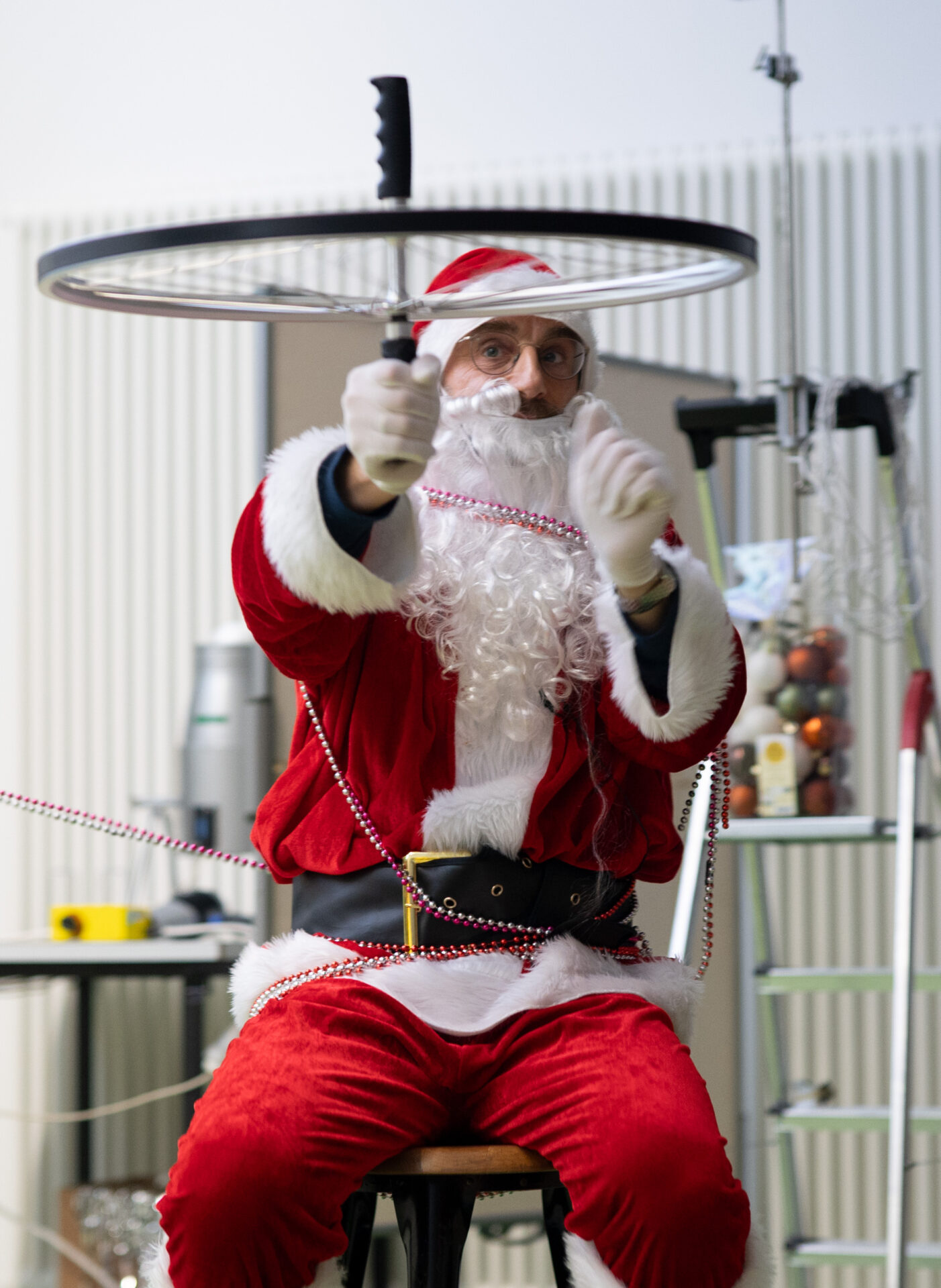 Beim Weihnachts-Zauber der Hochschule Coburg sitzt eine als Weihnachtsmann verkleidete Person auf einem Hocker und dreht ein Messer, das auf einem Fahrradrad balanciert, mit einer Trittleiter und verschiedenen Gegenständen im Hintergrund - eine experimentelle Vorlesung im festlichen Stil. at Hochschule Coburg