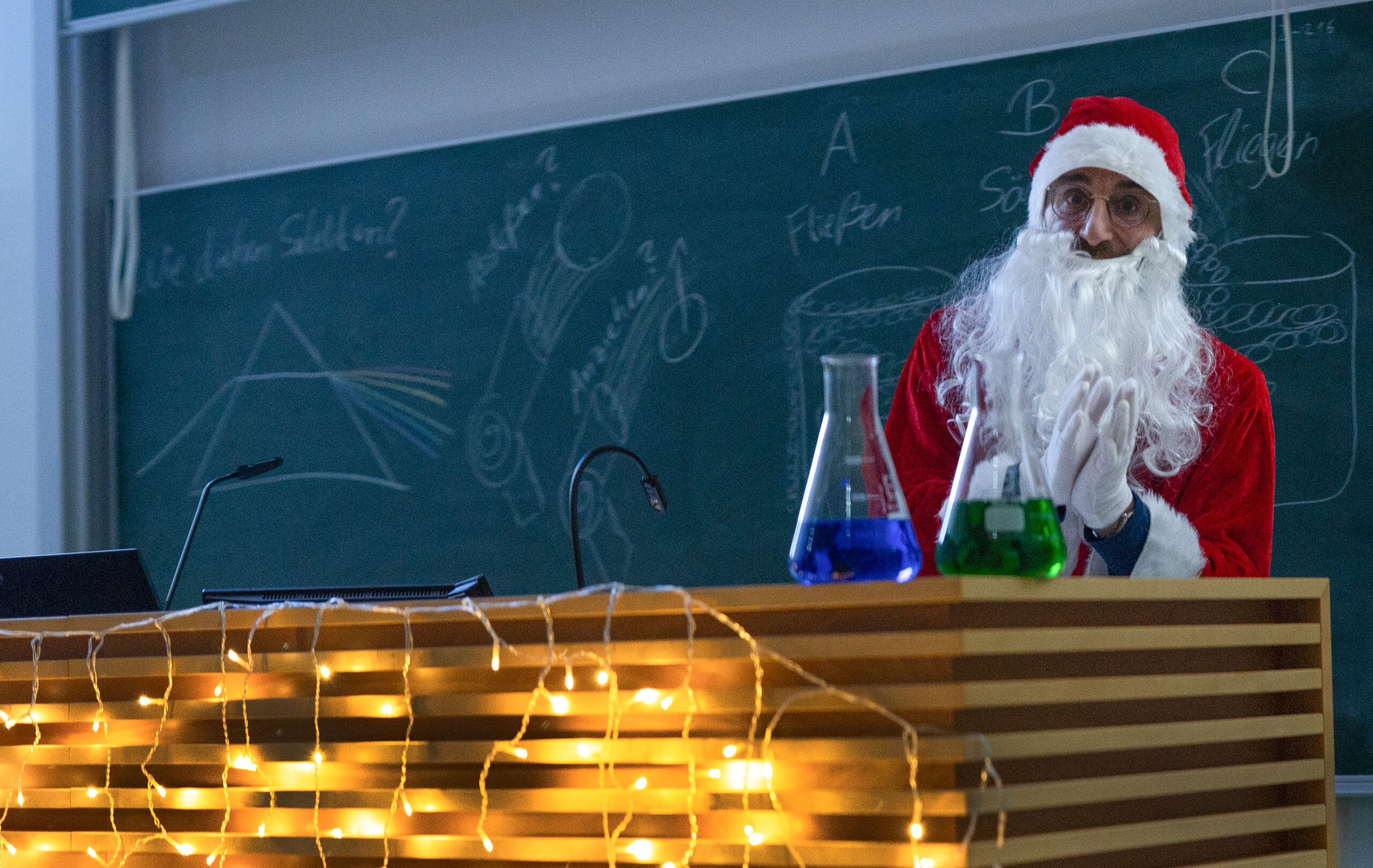 Eine als Weihnachtsmann verkleidete Person steht hinter einem mit Lichterketten und Laborkolben geschmückten Schreibtisch und bringt Weihnachts-Zauber in eine experimentelle Vorlesung an der Hochschule Coburg, vor einer Kreidetafel, die mit wissenschaftlichen Zeichnungen und Notizen gefüllt ist. at Hochschule Coburg