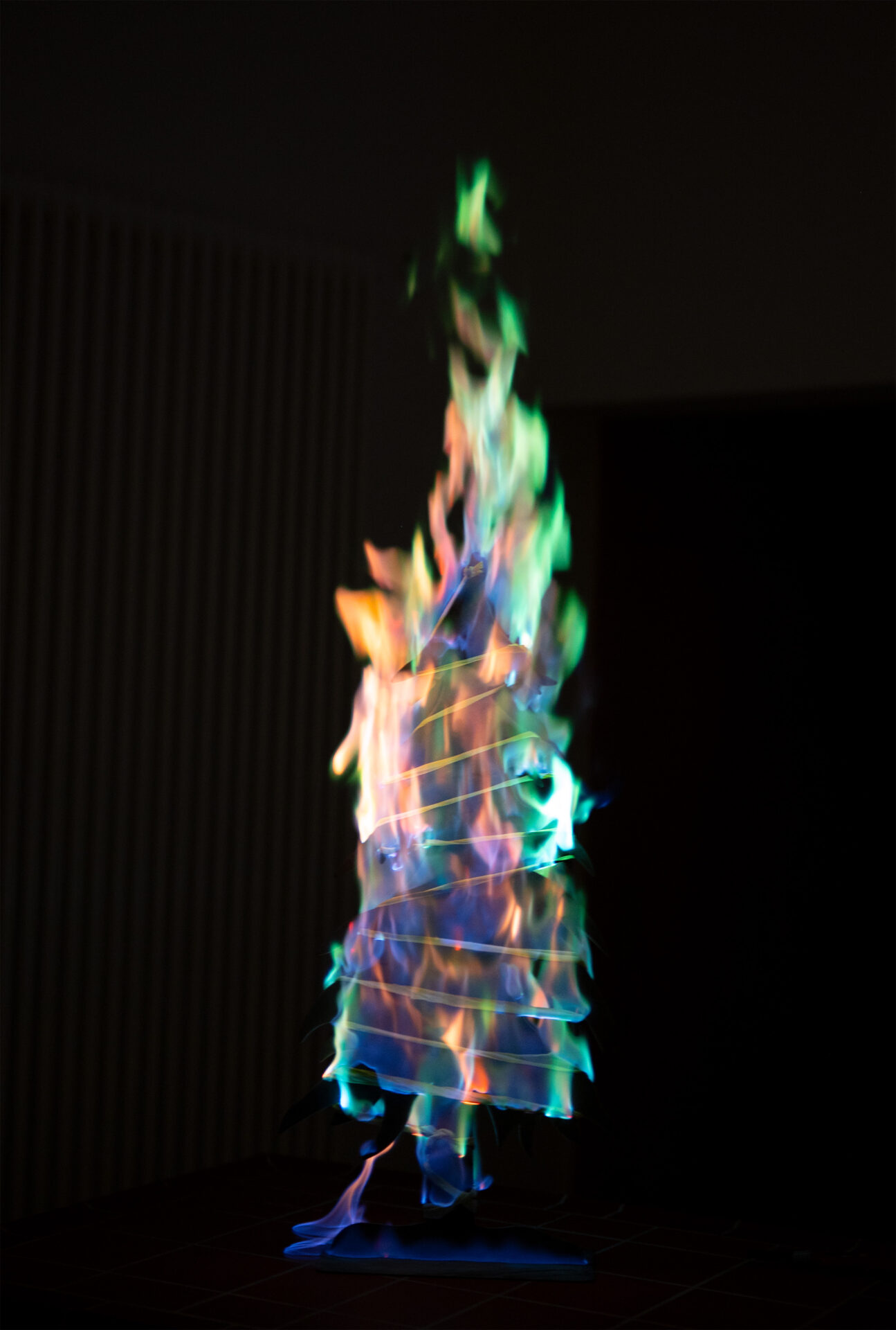 Ein Papierstapel brennt mit bunten Flammen in Grün-, Blau-, Lila- und Orangetönen vor dunklem Hintergrund - eine Reminiszenz an die experimentelle Vorlesung der Hochschule Coburg und ihren schillernden Weihnachts-Zauber. at Hochschule Coburg