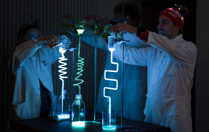 Drei Personen in Laborkitteln an der Hochschule Coburg gießen leuchtende Flüssigkeiten in spiralförmige Röhren, die hell leuchten und einen Weihnachts-Zauber-Effekt erzeugen. Eine Person, die eine Weihnachtsmütze mit blinkenden Lichtern trägt, verleiht dieser experimentellen Vorlesung eine festliche Note. at Hochschule Coburg