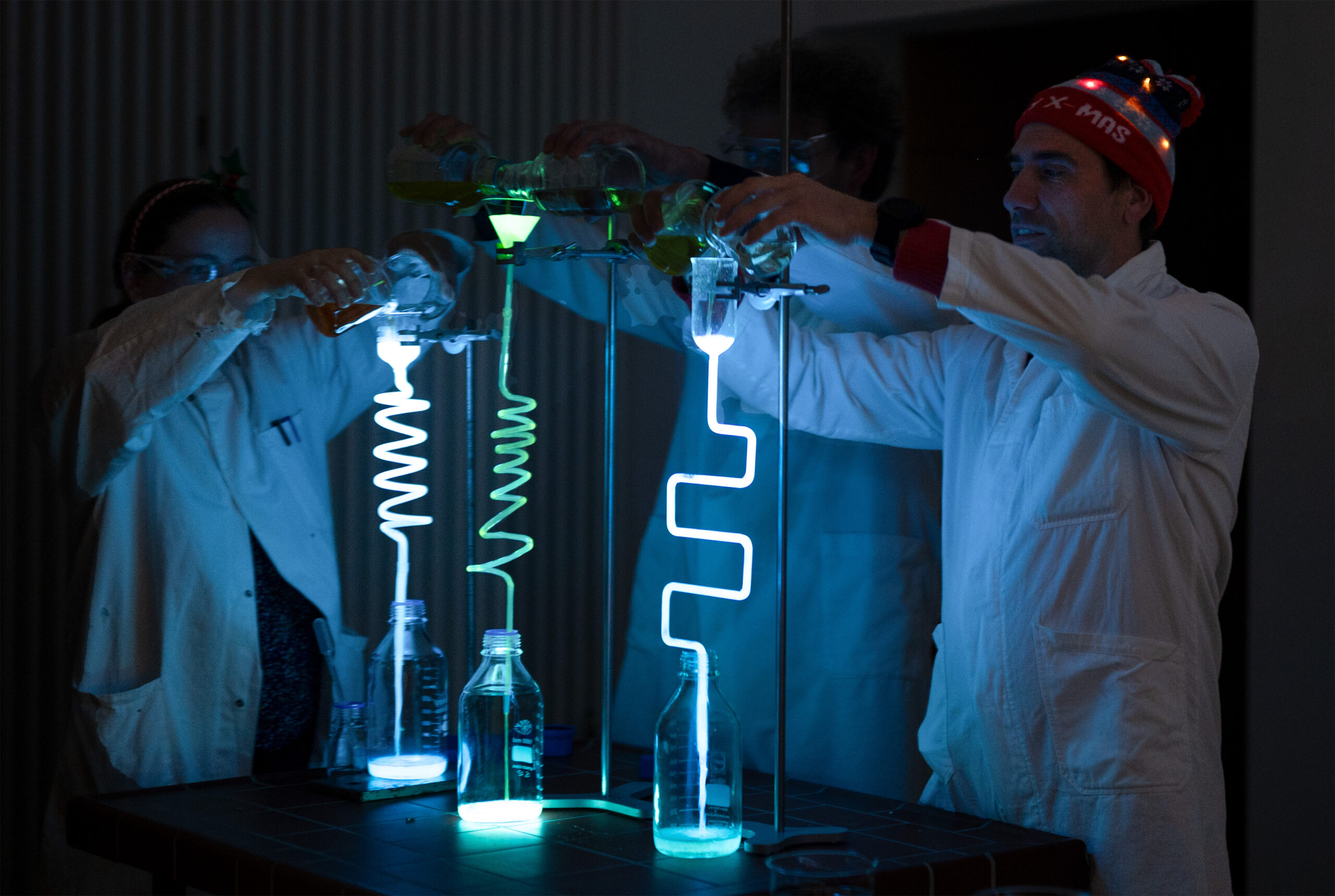 Drei Personen in Laborkitteln an der Hochschule Coburg gießen leuchtende Flüssigkeiten in spiralförmige Röhren, die hell leuchten und einen Weihnachts-Zauber-Effekt erzeugen. Eine Person, die eine Weihnachtsmütze mit blinkenden Lichtern trägt, verleiht dieser experimentellen Vorlesung eine festliche Note. at Hochschule Coburg