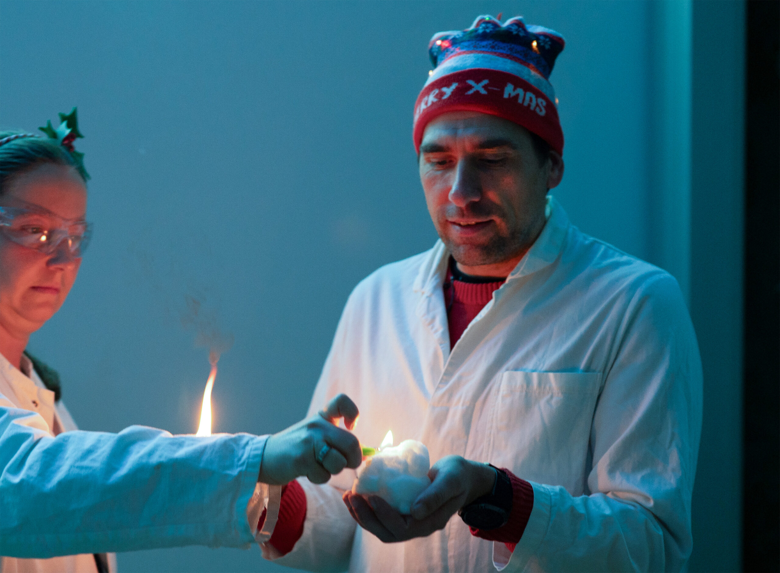 Zwei Menschen in Laborkitteln, einer mit rotem "Merry X-mas"-Hut, halten ein weißes Objekt, während der andere es mit einer Flamme anzündet. Blaues Licht sorgt für wissenschaftliche Stimmung - es ist eine Experimentelle Vorlesung an der Hochschule Coburg, die puren Weihnachts-Zauber einfängt. at Hochschule Coburg