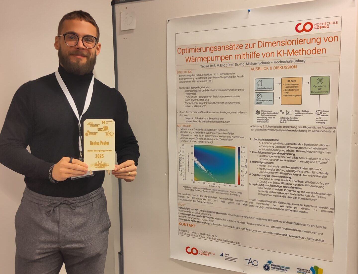 Ein lächelnder Mann mit Brille und Bart hält eine Best-Poster-Award-Trophäe in der Hand, während er neben seinem deutschen Forschungsposter zur Optimierung von Wärmepumpen mit KI-Innovation steht. Das Poster ist auf einer Tafel in einem weißwandigen Raum ausgestellt. at Hochschule Coburg