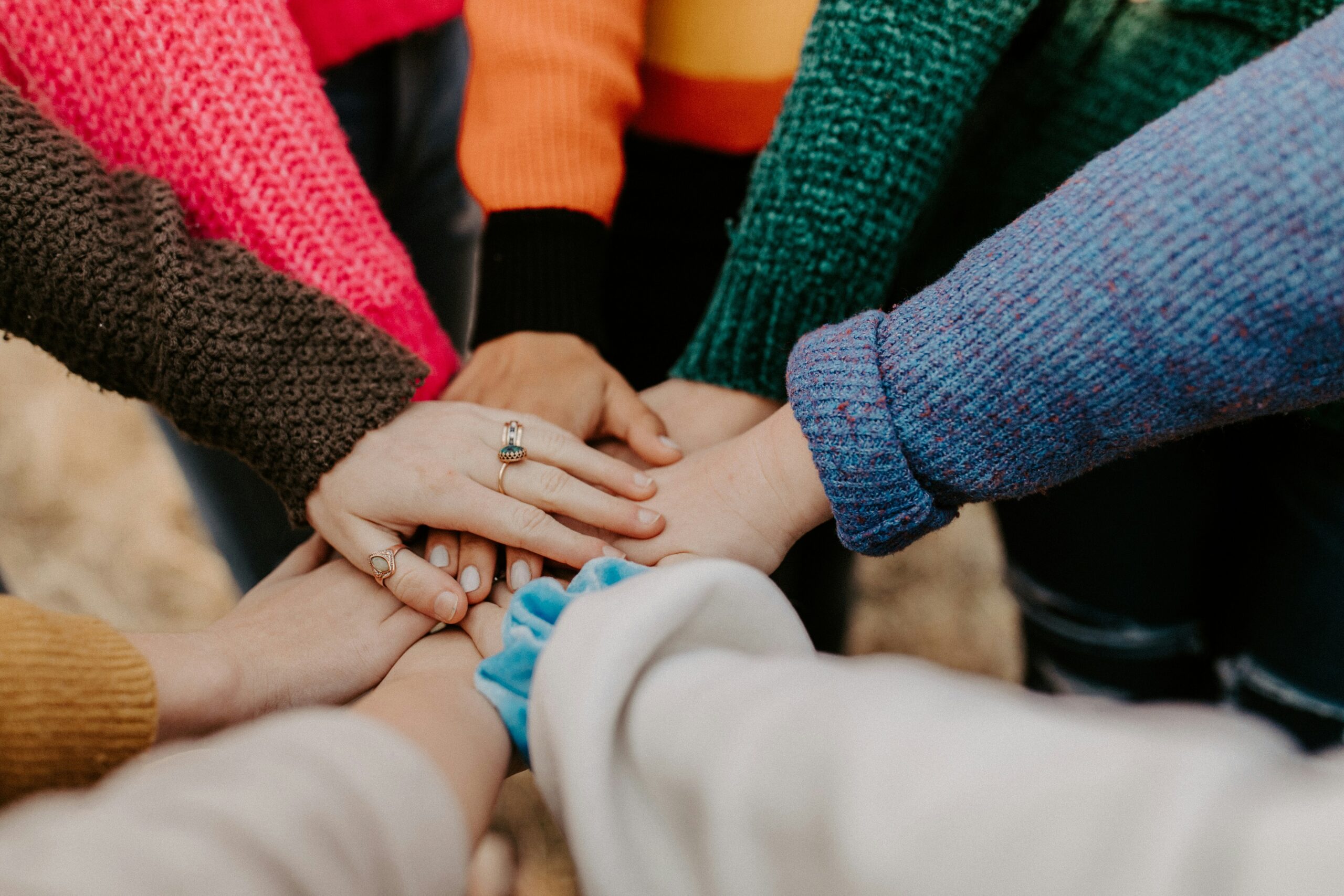 Eine Gruppe von Menschen, die in einem Kreis stehen, legen ihre Hände in der Mitte zusammen und tragen bunte Pullover, die Vielfalt, Menschlichkeit, Einigkeit und Teamwork symbolisieren. at Hochschule Coburg