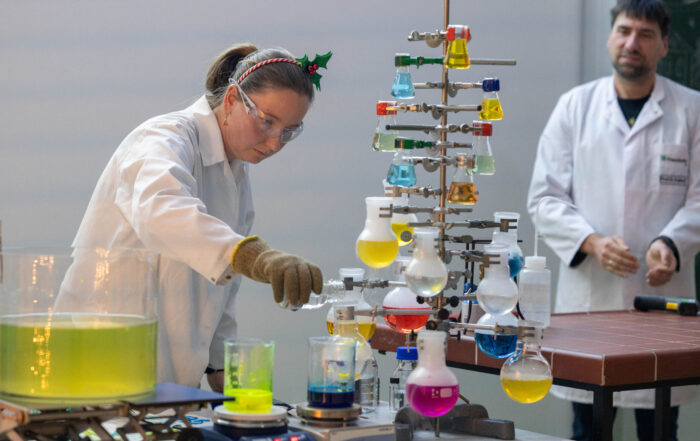 Bei einer Weihnachtsvorlesung im Labor der Hochschule gießt eine Frau mit Schutzbrille und weihnachtlichem Stirnband eine Flüssigkeit in einen Kolben, der von mit Chemikalien gefüllten Gläsern umgeben ist, die wie ein Weihnachtsbaum angeordnet sind, während ein Mann im Laborkittel zusieht. at Hochschule Coburg