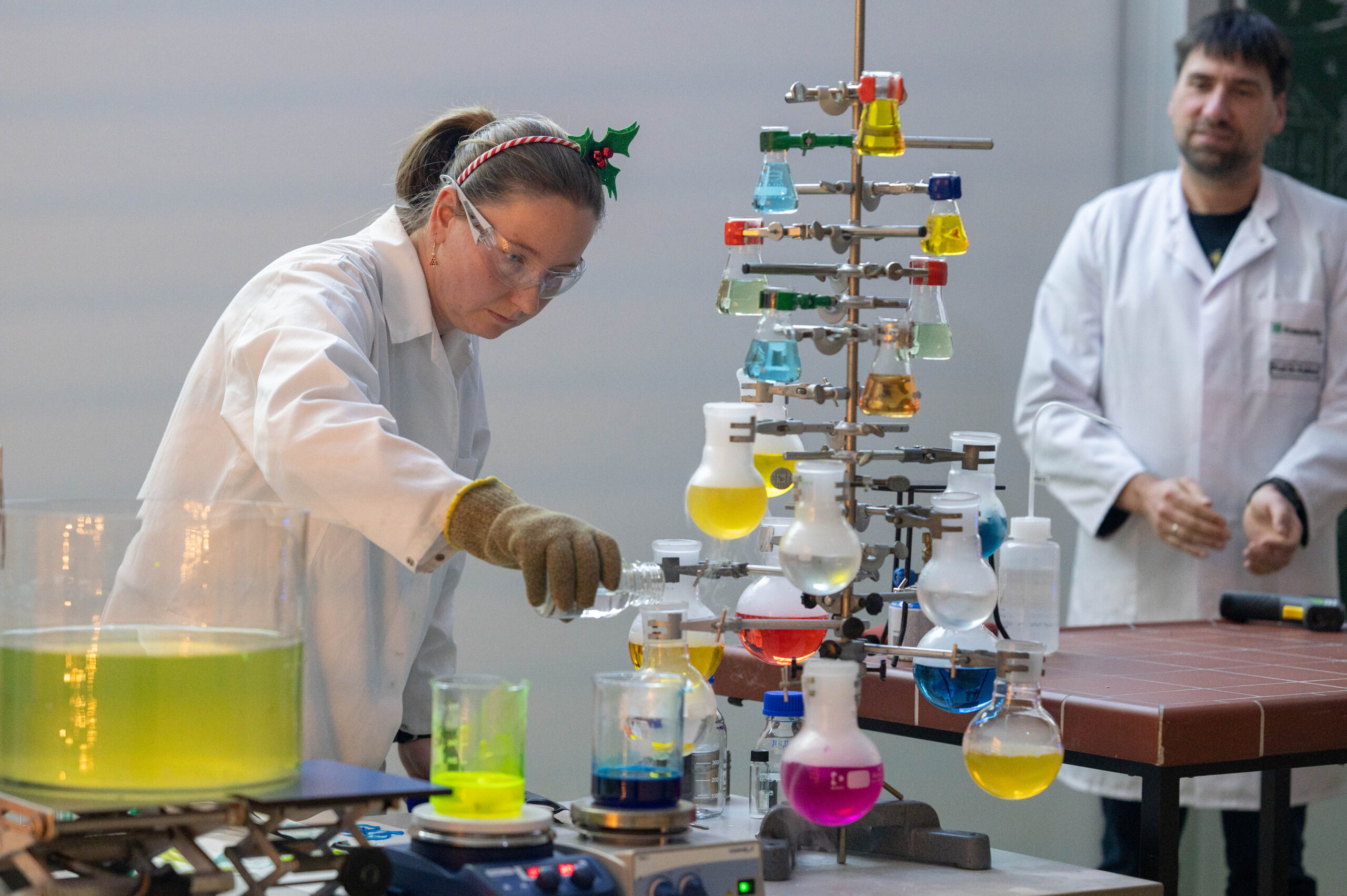 Bei einer Weihnachtsvorlesung im Labor der Hochschule gießt eine Frau mit Schutzbrille und weihnachtlichem Stirnband eine Flüssigkeit in einen Kolben, der von mit Chemikalien gefüllten Gläsern umgeben ist, die wie ein Weihnachtsbaum angeordnet sind, während ein Mann im Laborkittel zusieht. at Hochschule Coburg