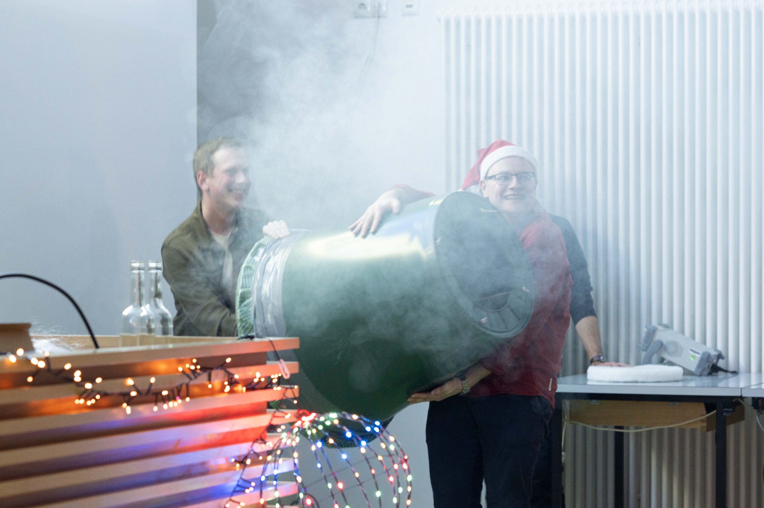 Bei einer festlichen Indoor-Veranstaltung halten zwei Personen - eine davon mit Weihnachtsmannmütze - einen großen, rauchgrünen Behälter als Teil der Weihnachtsvorlesung von Wunderbare Wissenschaft. Im Vordergrund glitzern Lichter und Dekorationen. at Hochschule Coburg