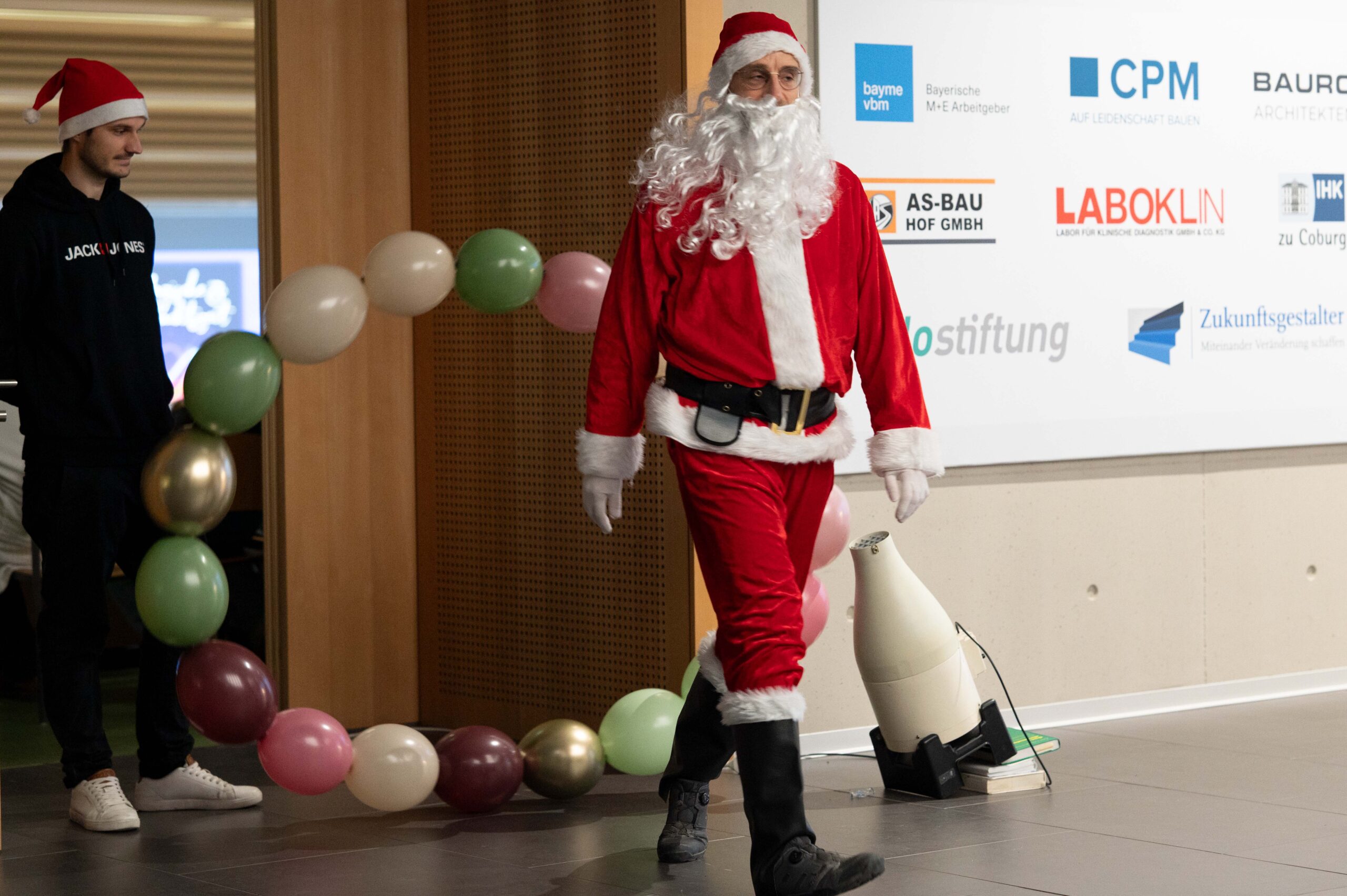 Bei einer Weihnachtsvorlesung geht eine als Weihnachtsmann verkleidete Person in der Nähe von Luftballondekorationen durch den Innenraum, während eine andere Person in einem schwarzen Kapuzenpulli in der Nähe steht. An der Wand im Hintergrund sind Firmenlogos und Hinweise auf die Hochschule Wissenschaft zu sehen. at Hochschule Coburg
