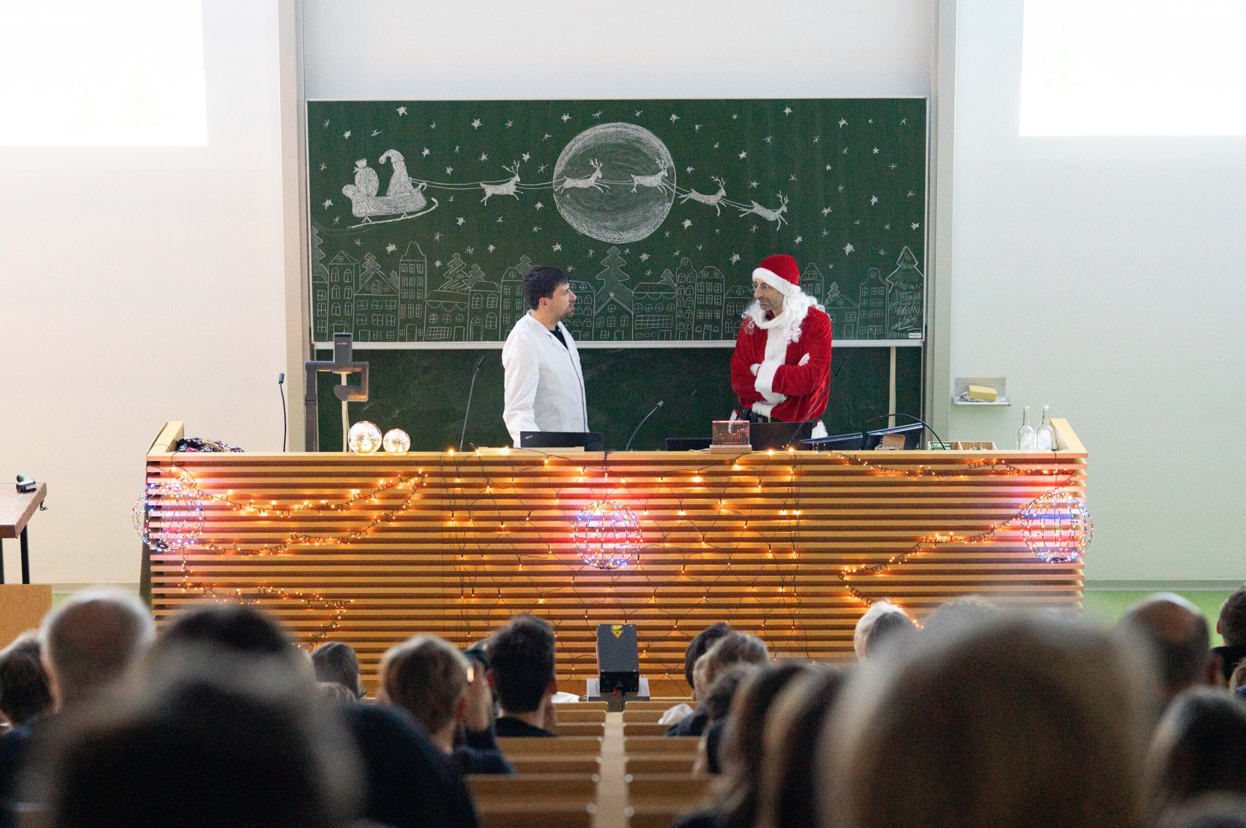 In einem festlich beleuchteten Klassenzimmer der Hochschule halten eine Person in einem weißen Laborkittel und ein weiterer als Weihnachtsmann eine Weihnachtsvorlesung von einem geschmückten Rednerpult aus, hinter dem der Schlitten des Weihnachtsmanns auf einer Kreidetafel zu sehen ist. at Hochschule Coburg
