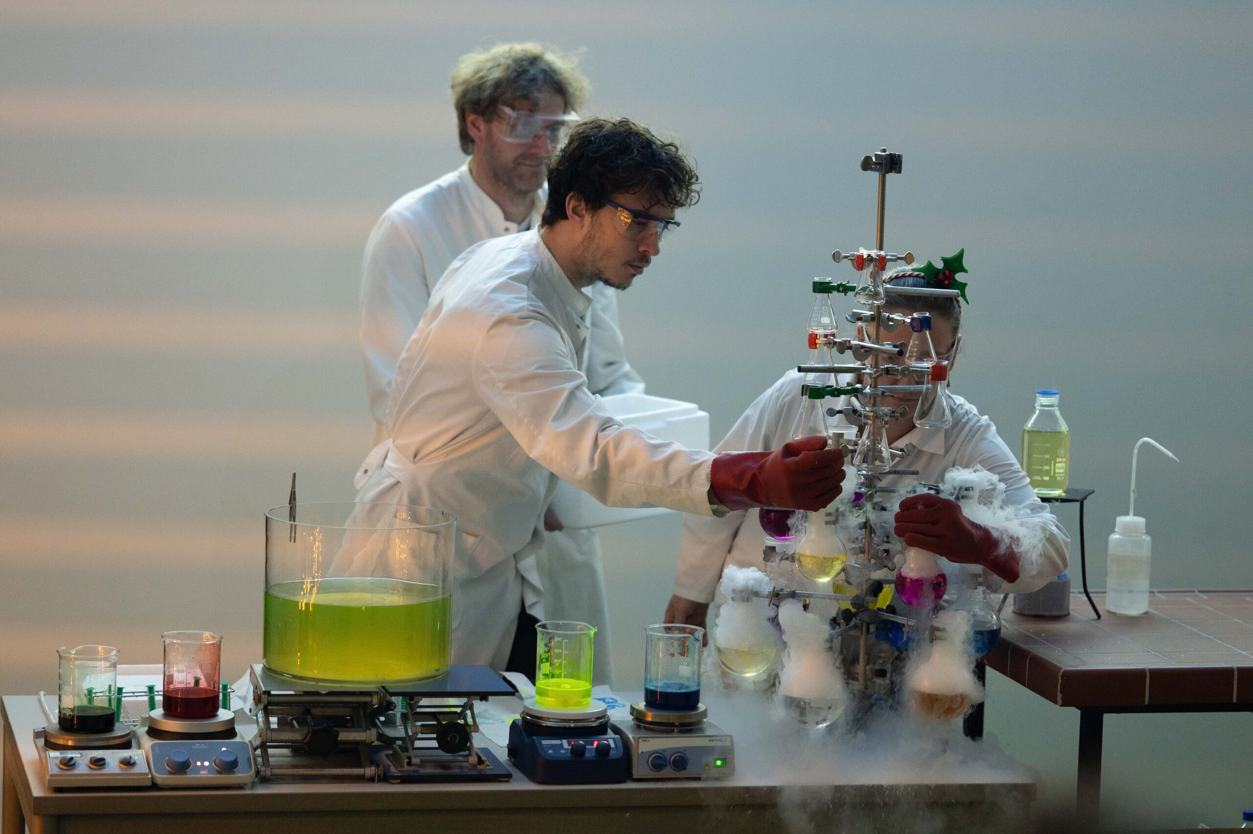 Drei Wissenschaftler in Laborkitteln und mit Schutzbrillen führen eine Weihnachtsvorlesung an der Hochschule durch. Sie experimentieren mit bunten Flüssigkeiten und Trockeneis und erzeugen Nebel auf einem Tisch, der mit Bechern und wissenschaftlichen Geräten gefüllt ist. at Hochschule Coburg
