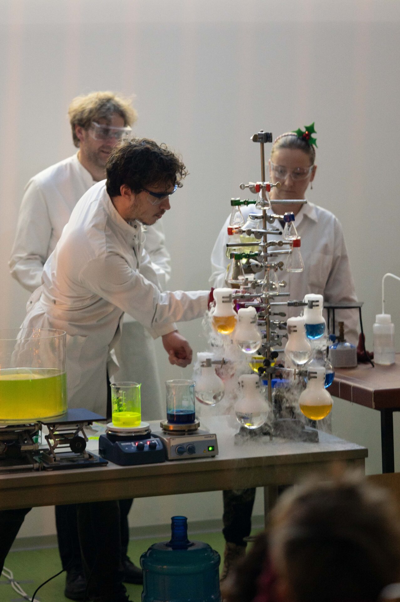 Drei Personen in Laborkitteln und Schutzbrillen führen in einem Labor der Hochschule ein chemisches Experiment mit bunten Flüssigkeiten in Glaskolben durch, das an eine festliche Weihnachtsvorlesung erinnert. at Hochschule Coburg