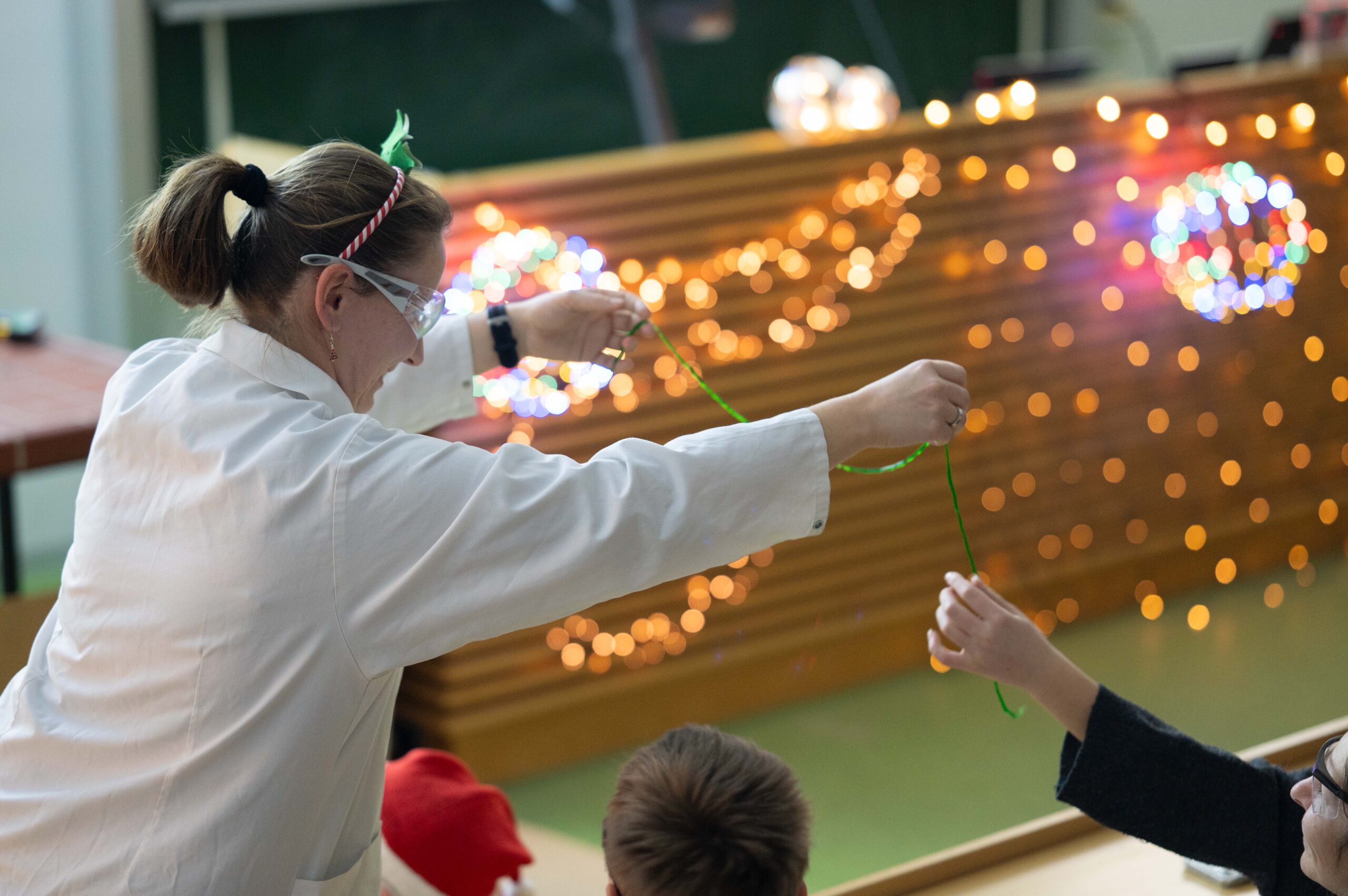 Eine Person in einem weißen Laborkittel führt Kindern in einem mit bunten Lichterketten dekorierten Klassenzimmer ein wissenschaftliches Experiment vor. Die Person hält einen grünen Gegenstand in der Hand, den ein Kind anfassen möchte. at Hochschule Coburg