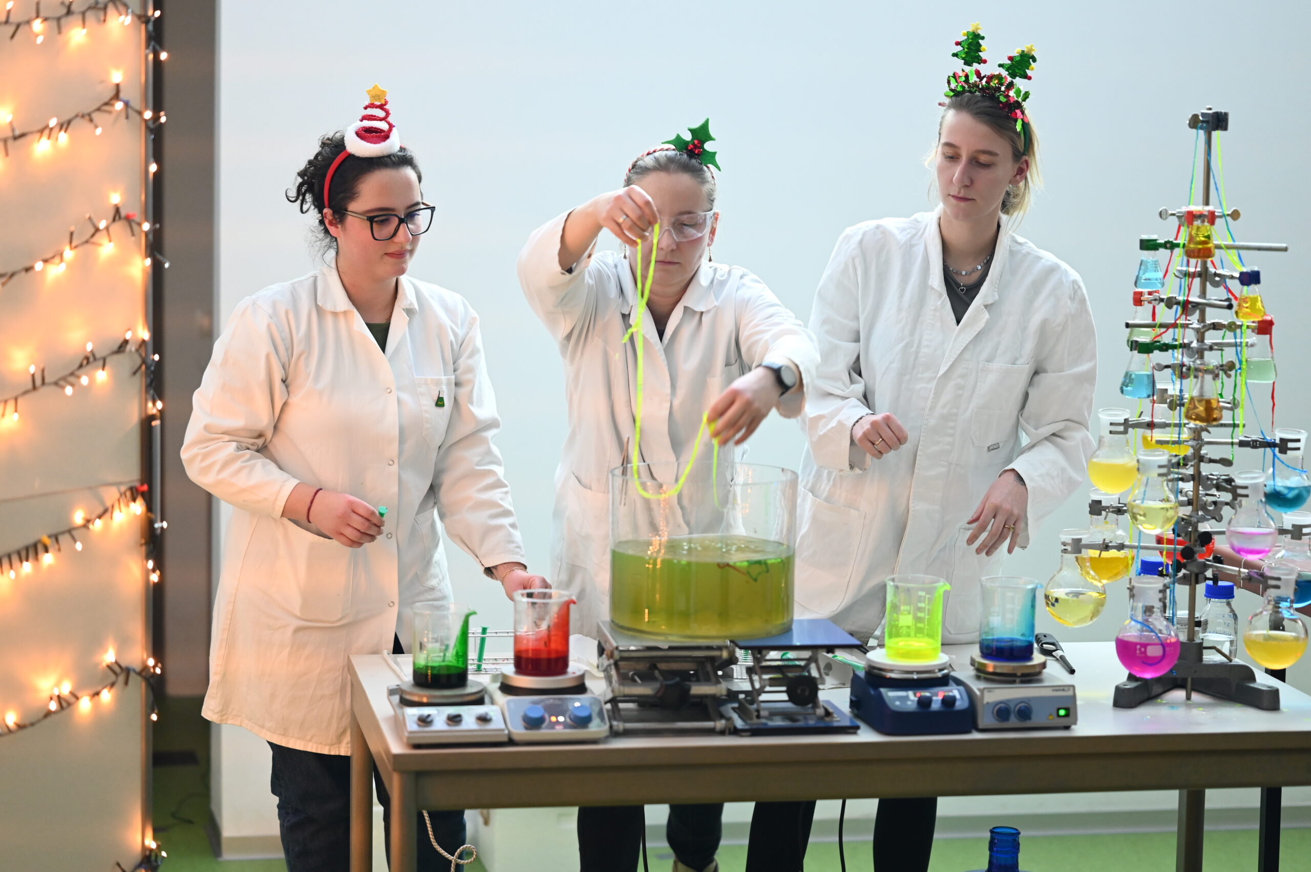 Drei Personen in Laborkitteln und mit festlichen Hüten führen ein buntes wissenschaftliches Experiment mit Bechern und Flüssigkeiten an einem Tisch durch. Festliche Lichter und Dekorationen erhellen diese lebhafte Weihnachtsvorlesung an der Hochschule. at Hochschule Coburg