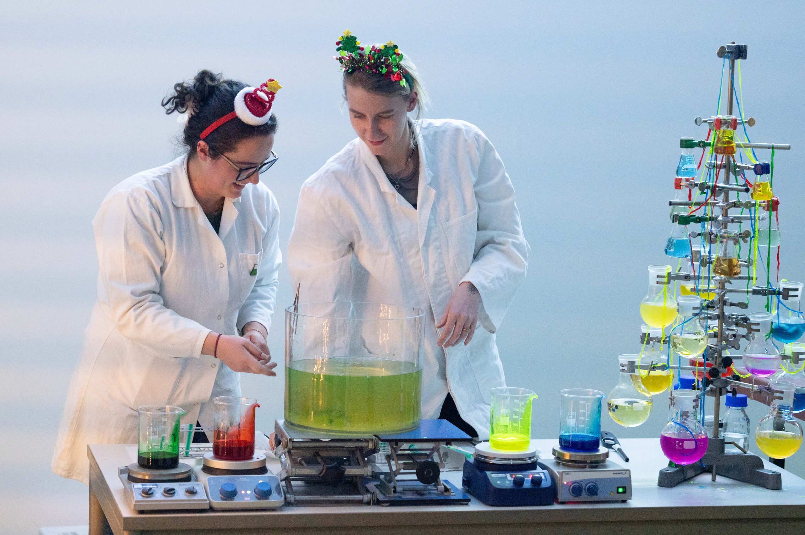 Zwei Frauen in Laborkitteln, die festliche Stirnbänder tragen, führen ein wissenschaftliches Experiment mit bunten Flüssigkeiten in Bechergläsern und einem großen Behälter auf einem Tisch durch, der neben einem Regal mit leuchtenden Lösungen steht und die Stimmung einer Weihnachtsvorlesung an der Hochschule einfängt. at Hochschule Coburg