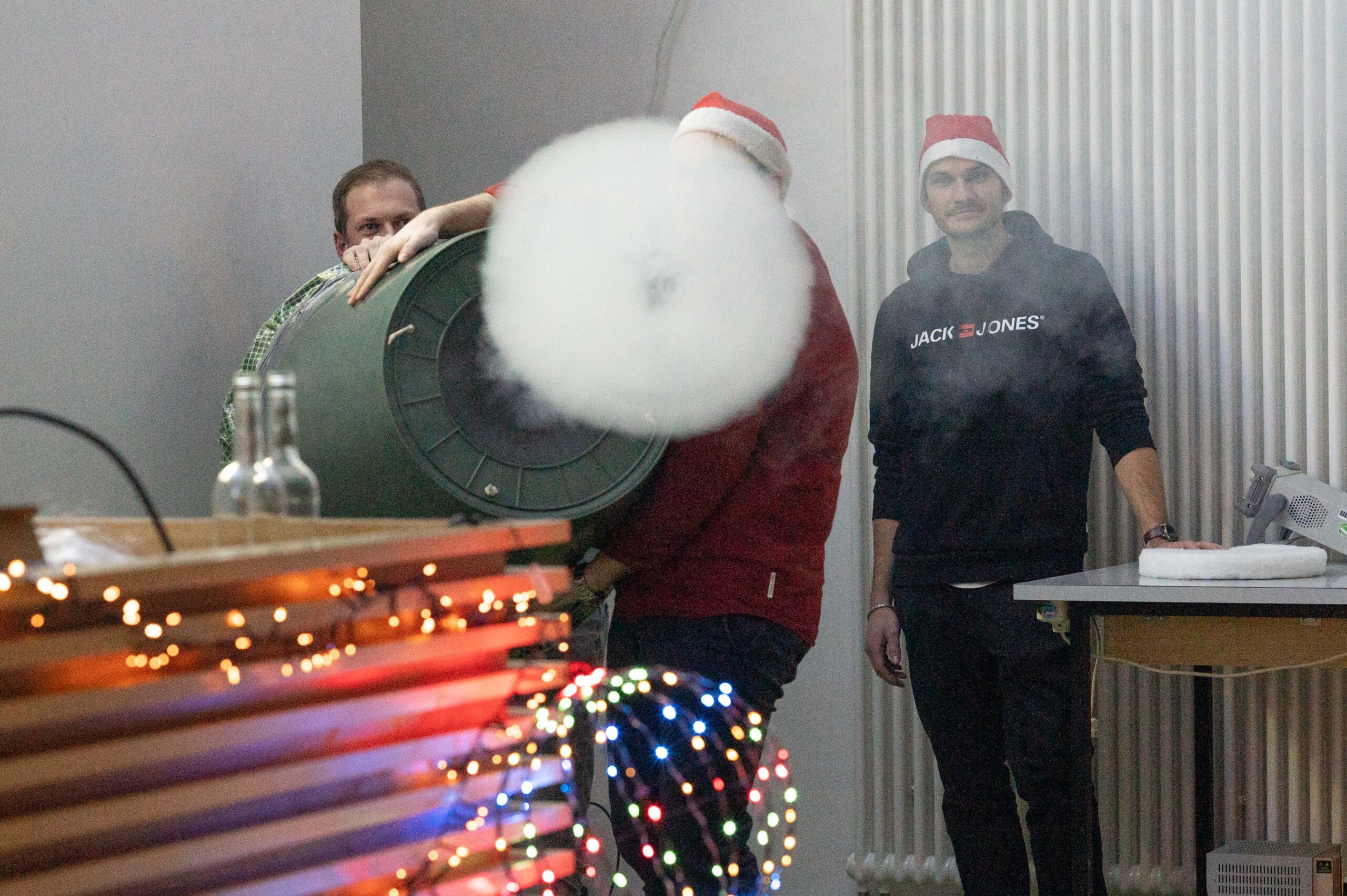 Drei Personen in einem Haus, zwei mit Weihnachtsmannmützen, feiern bei einer Weihnachtsvorlesung. Einer feuert eine große Luftkanone ab, die einen Rauchring durch Lichterketten und Weihnachtsdekoration schickt. In der festlichen Umgebung der Hochschule sind wissenschaftliche Geräte und eine Flasche zu sehen. at Hochschule Coburg