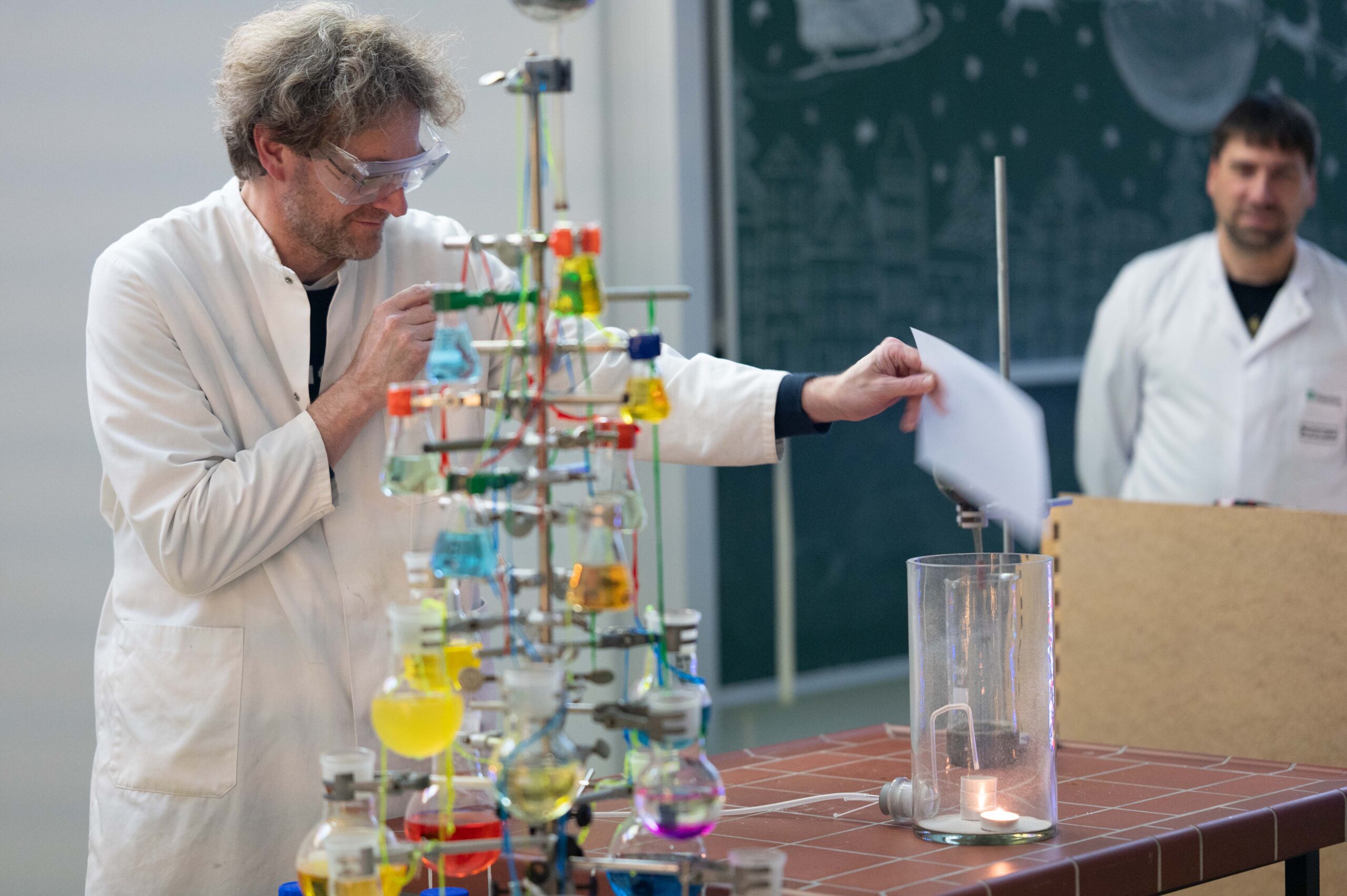 Während einer Weihnachtsvorlesung an der Hochschule hält ein Wissenschaftler in einem weißen Laborkittel und mit Schutzbrille ein Blatt Papier über einen hohen Glaszylinder mit einer Flamme darin. Bunte wissenschaftliche Geräte stehen in der Nähe, während eine andere Person im Hintergrund beobachtet. at Hochschule Coburg