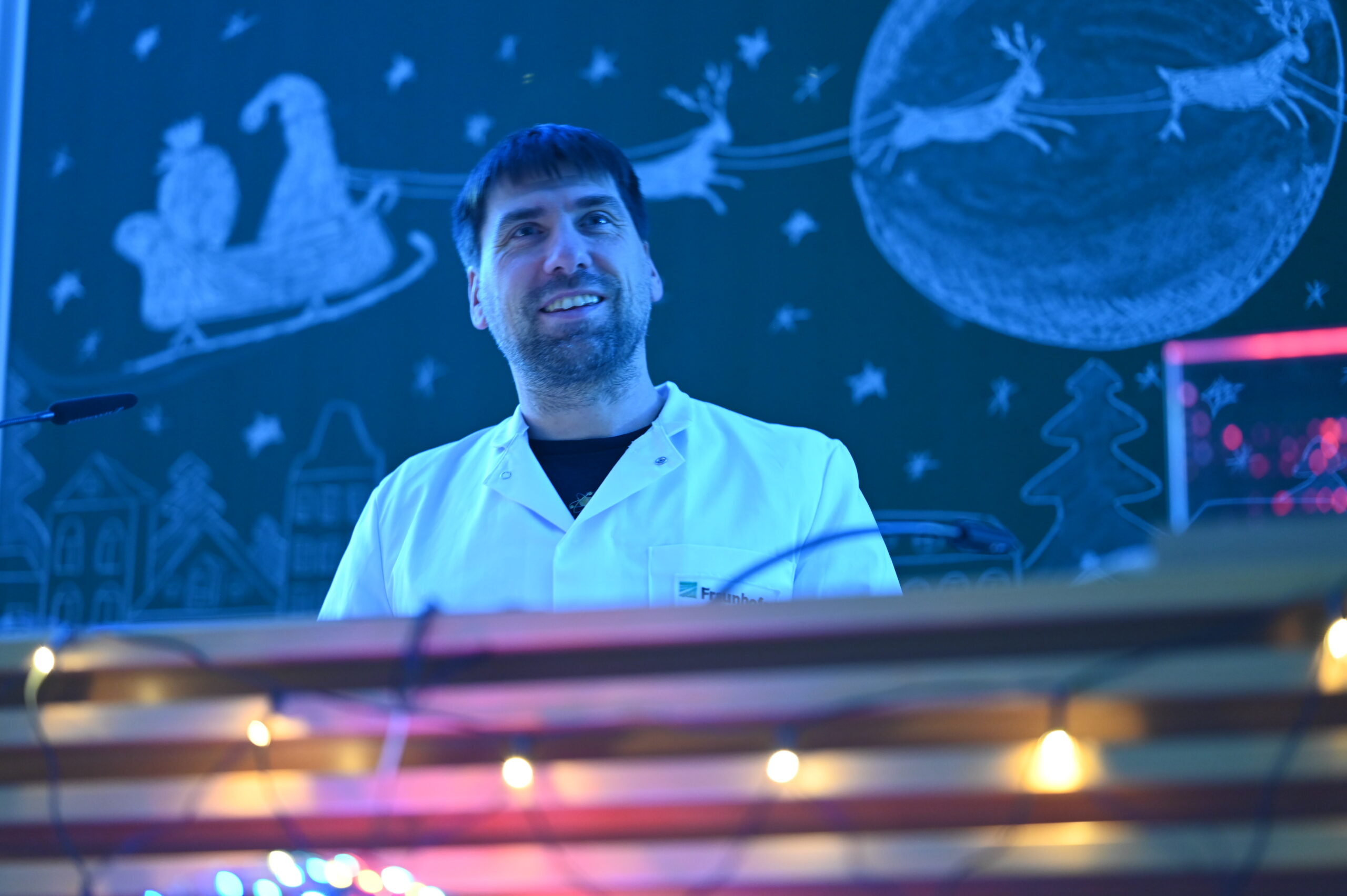 Ein Mann in einem weißen Laborkittel steht lächelnd vor einer Kreidezeichnung des Schlittens und der Rentiere des Weihnachtsmanns, mit festlichen Lichtern im Vordergrund - und fängt den Geist der Wissenschaft bei einer fröhlichen Weihnachtsvorlesung an der Hochschule ein. at Hochschule Coburg