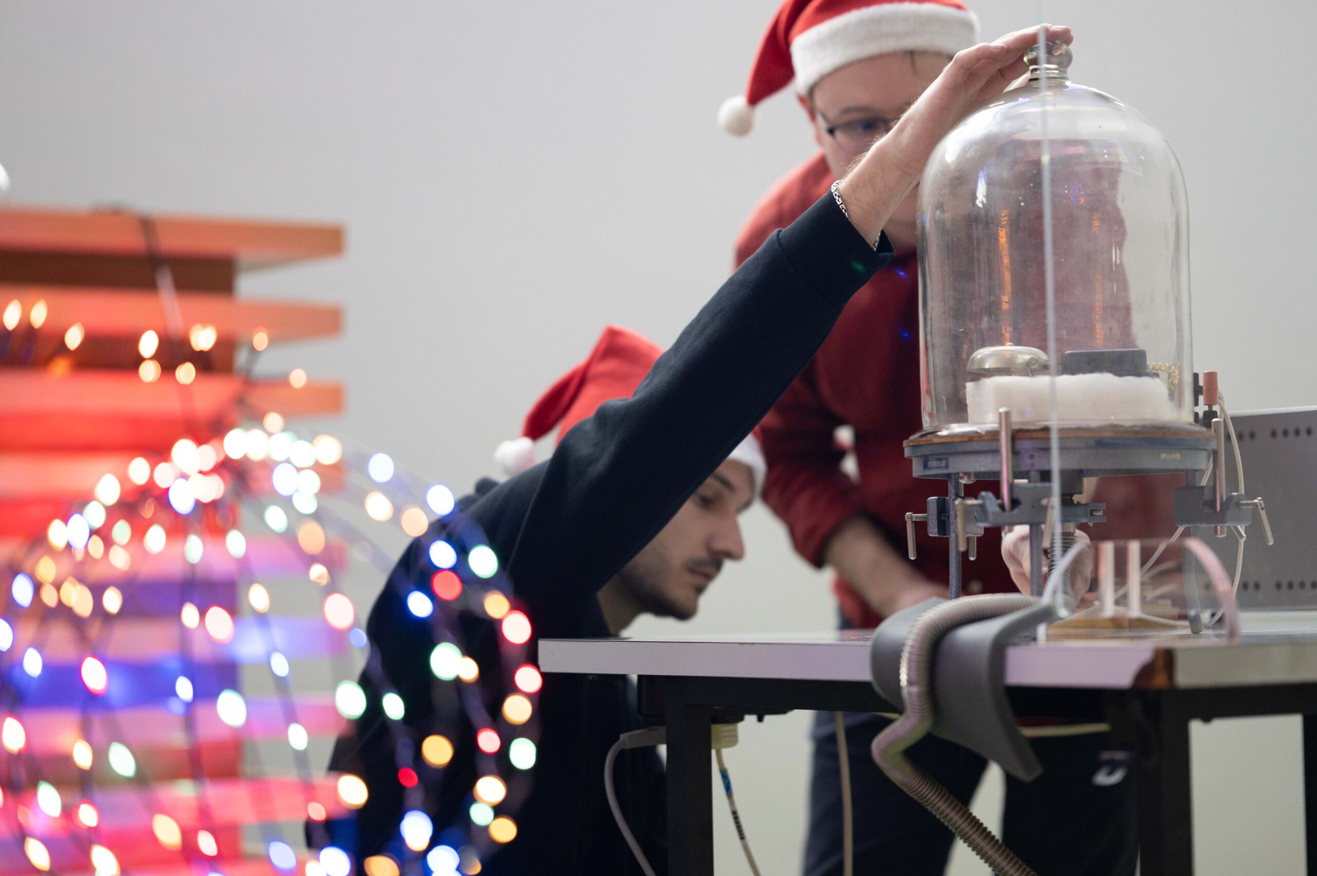 Zwei Personen mit Weihnachtsmannmützen führen ein wissenschaftliches Experiment mit einem Glasbehälter auf einem Tisch durch. Im Vordergrund sind unscharfe Lichter zu sehen, die eine festliche Weihnachtsvorlesungsatmosphäre schaffen. at Hochschule Coburg