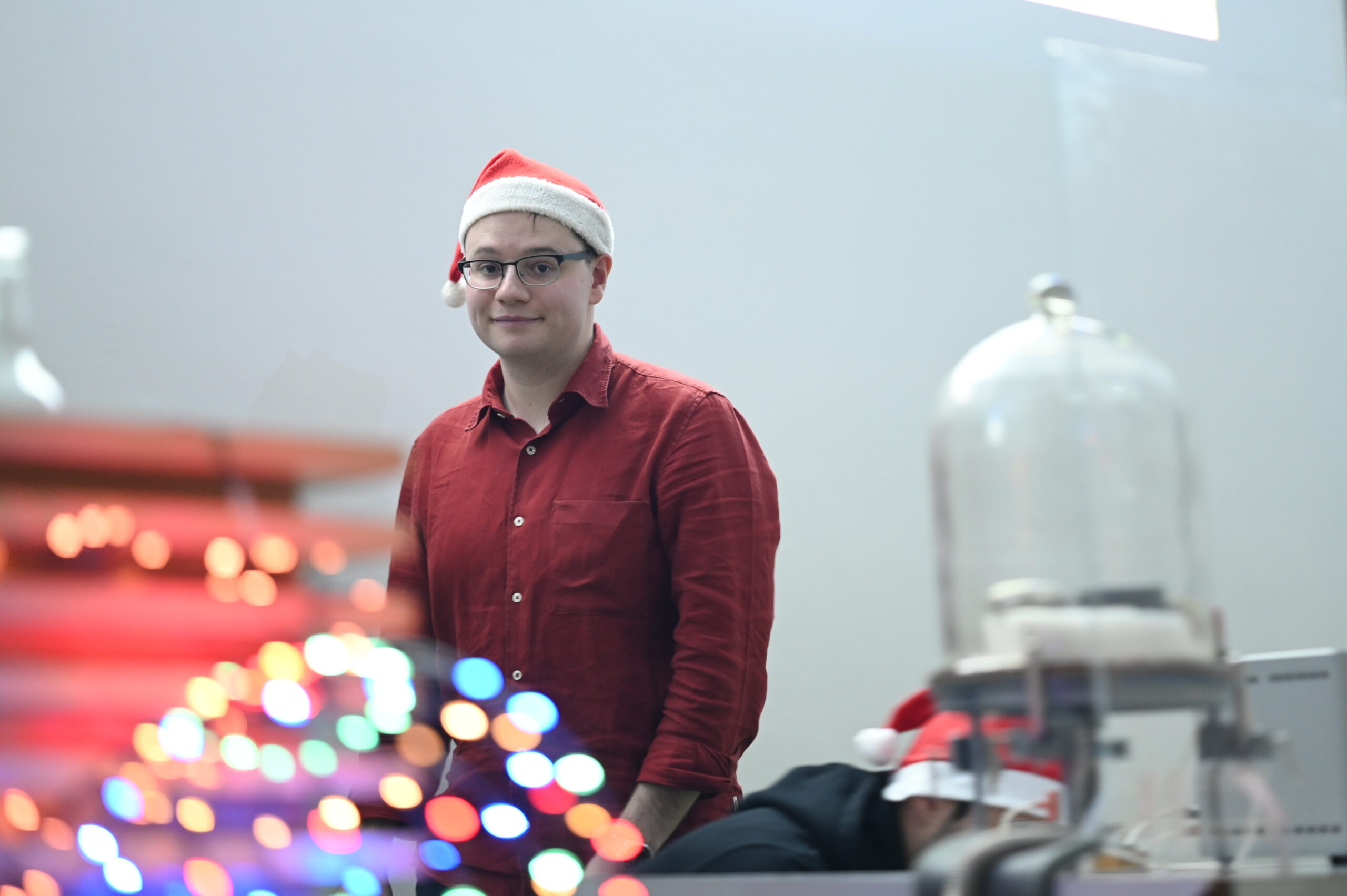 Eine Person mit Brille und Weihnachtsmannmütze steht mit einem roten Hemd bekleidet in einem Innenraum einer Hochschule. Im Vordergrund deuten bunte Lichterketten und unscharfe Dekorationen auf eine festliche Weihnachtsvorlesung hin. Eine weitere Person mit Weihnachtsmannmütze ist teilweise sichtbar, sitzend. at Hochschule Coburg