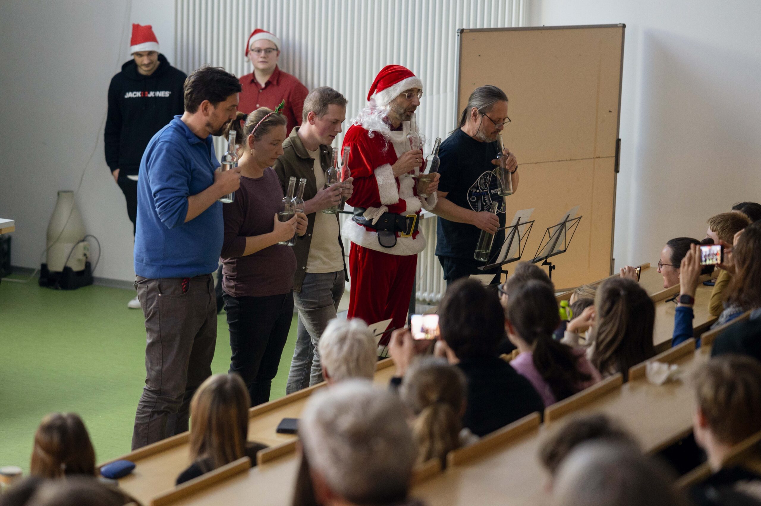 Während einer Weihnachtsvorlesung an der Hochschule spielt eine Gruppe Erwachsener - darunter einer als Weihnachtsmann - mit Gläsern Musik für ein Publikum in einem Klassenzimmer. Mehrere Personen nehmen die Aufführung auf, während andere mit Weihnachtsmannmützen von hinten zusehen. at Hochschule Coburg