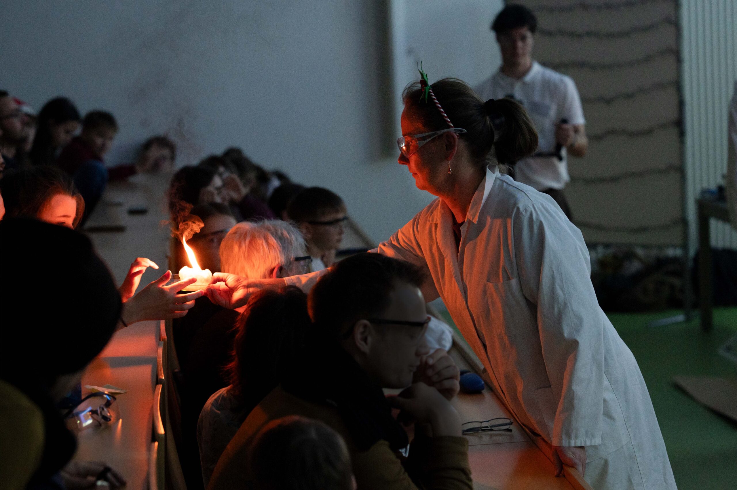 Während einer Weihnachtsvorlesung an der Hochschule hält eine Frau im Laborkittel und mit Schutzbrille einen brennenden Gegenstand vor ein Publikum aus Kindern und Erwachsenen und führt ein spannendes wissenschaftliches Experiment im abgedunkelten Klassenzimmer vor. at Hochschule Coburg