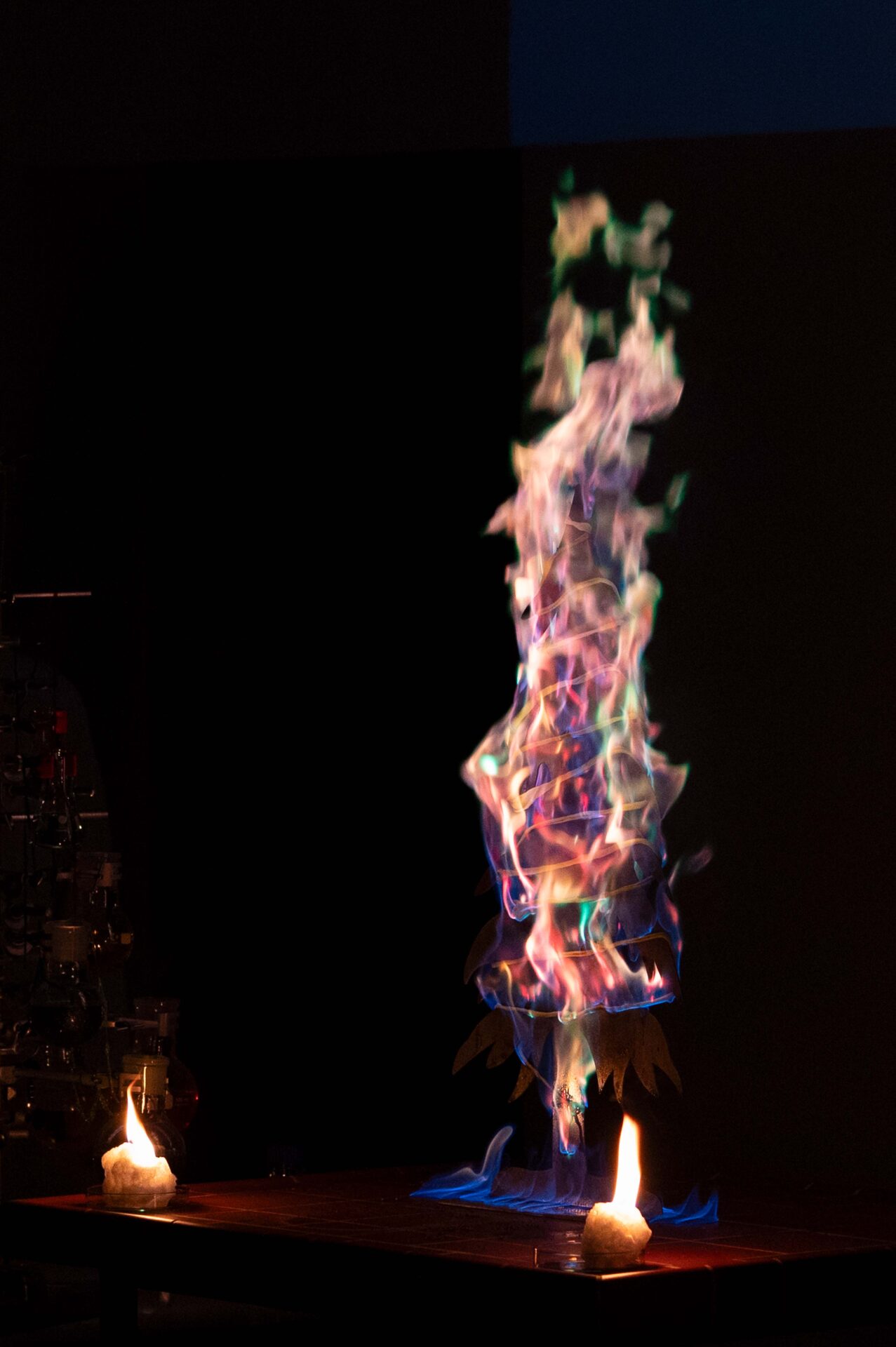 Eine leuchtende, mehrfarbige Flamme erhebt sich in einer gewundenen Form aus einer dunklen Oberfläche und schafft eine dramatische Darstellung - perfekt für eine Wunderbare Wissenschaft Weihnachtsvorlesung an der Hochschule - während zwei kleinere gelbe Flammen auf beiden Seiten vor einem schwarzen Hintergrund brennen. at Hochschule Coburg