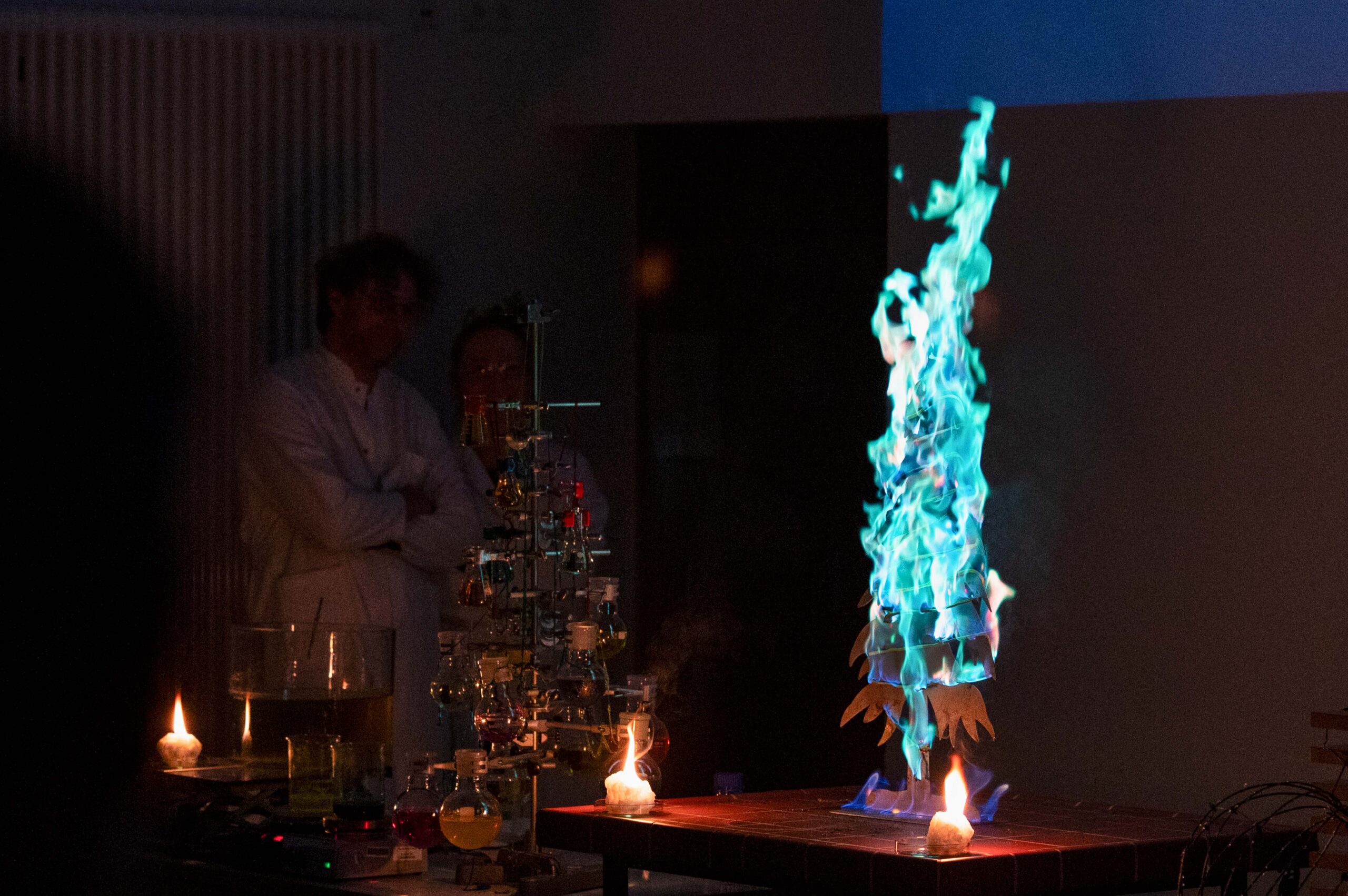 Zwei Personen in Laborkitteln beobachten in der Weihnachtsvorlesung der Hochschule ein Chemieexperiment, bei dem in einem schwach beleuchteten Raum leuchtend blaue und grüne Flammen aus wissenschaftlichen Gläsern und Geräten aufsteigen und die Begeisterung für die Wissenschaft zum Ausdruck bringen. at Hochschule Coburg