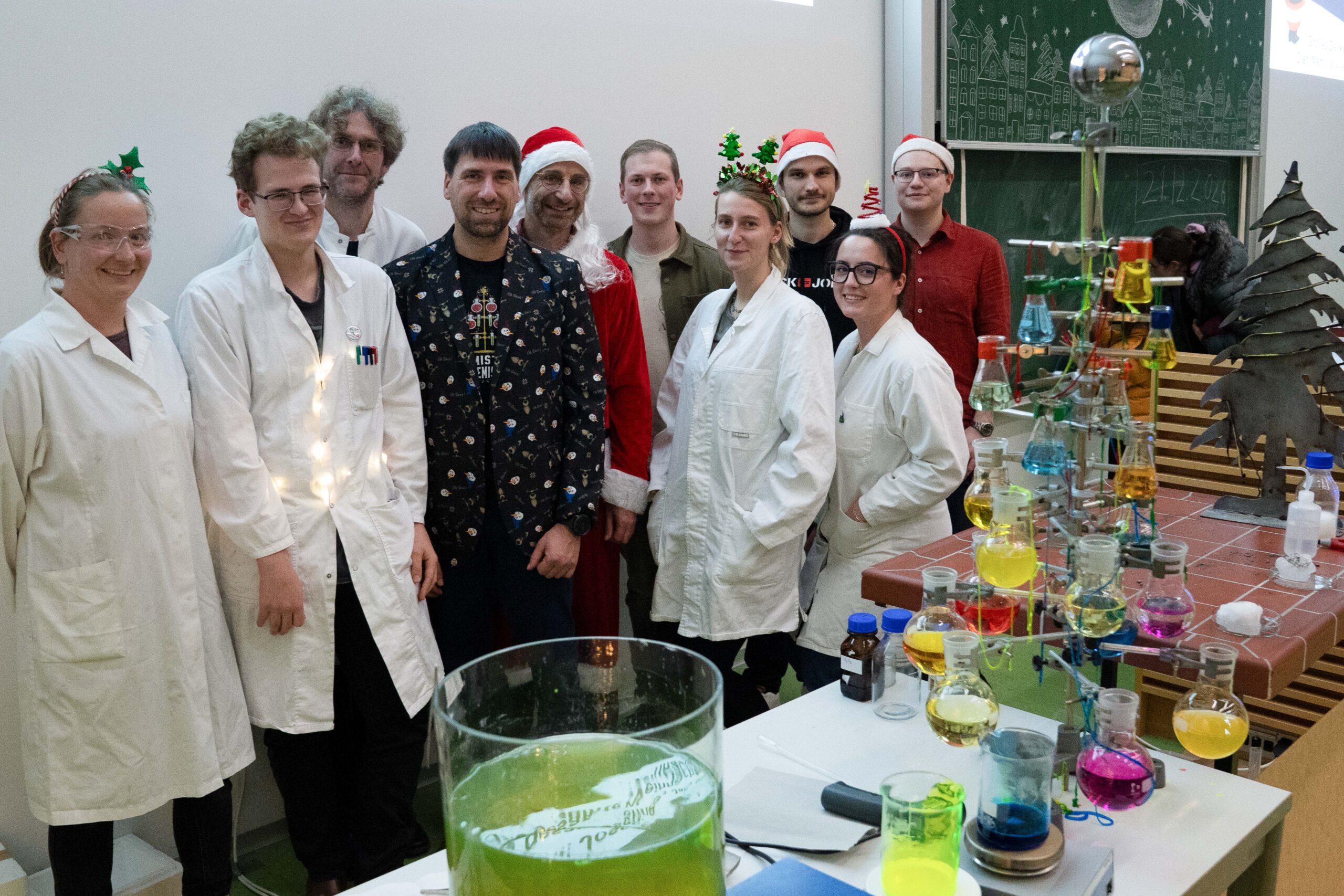 Eine Gruppe von Personen in Laborkitteln und mit festlichen Weihnachtsmützen posiert während der Weihnachtsvorlesung in einem naturwissenschaftlichen Klassenzimmer der Hochschule, mit bunten Bechern und Flaschen auf dem Tisch. Im Hintergrund sind eine Kreidetafel und eine Baumskulptur aus Metall zu sehen. at Hochschule Coburg