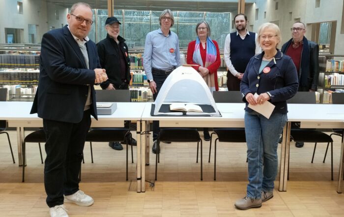 Eine Gruppe von sieben Erwachsenen steht lächelnd um eine Vitrine herum, in der sich ein aufgeschlagenes Buch befindet, in einer modernen Bibliothek mit großen Fenstern und Bücherregalen im Hintergrund. at Hochschule Coburg