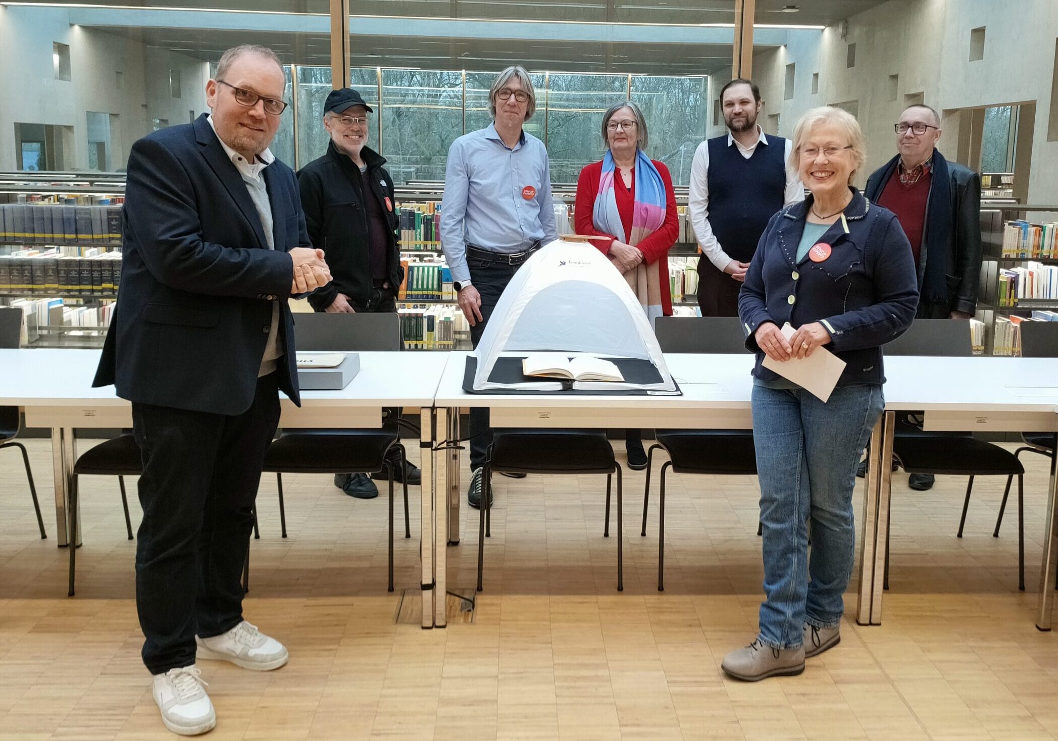 Eine Gruppe von sieben Erwachsenen steht lächelnd um eine Vitrine herum, in der sich ein aufgeschlagenes Buch befindet, in einer modernen Bibliothek mit großen Fenstern und Bücherregalen im Hintergrund. at Hochschule Coburg