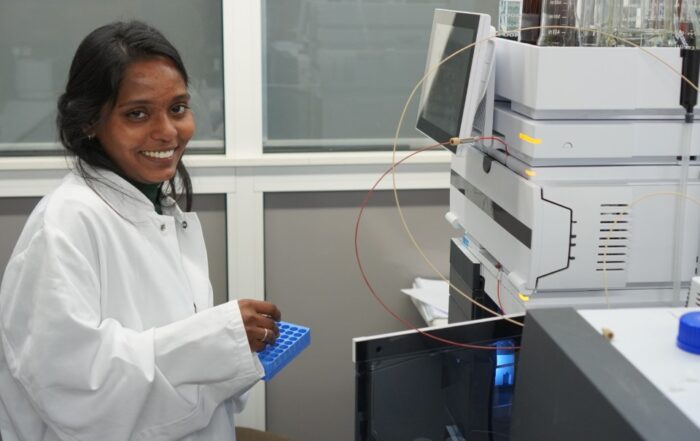 Eine Frau in einem weißen Laborkittel, möglicherweise eine indische Doktorandin, lächelt in die Kamera, während sie ein blaues Reagenzglasgestell in einem Coburger Labor hält. Sie steht neben wissenschaftlichen Geräten und einem Computermonitor und forscht vielleicht an Biomarkern. at Hochschule Coburg