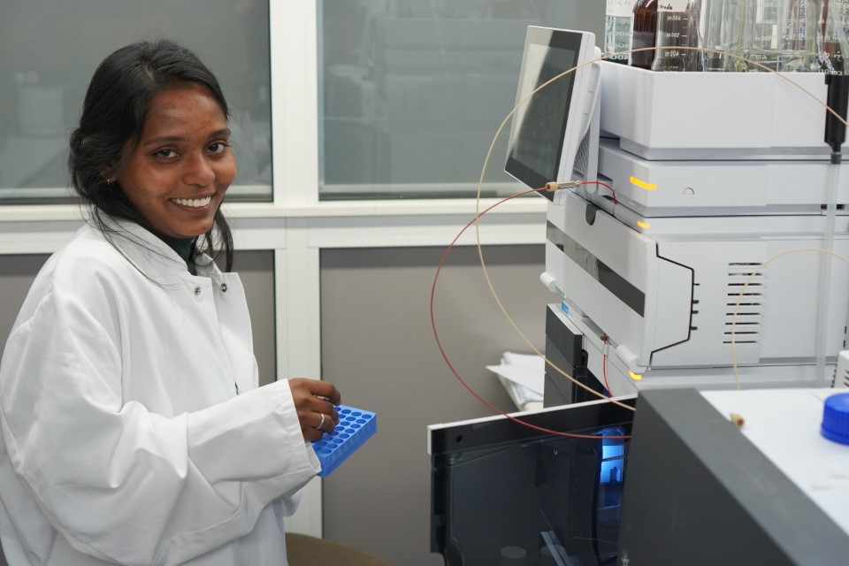 Eine Frau in einem weißen Laborkittel, möglicherweise eine indische Doktorandin, lächelt in die Kamera, während sie ein blaues Reagenzglasgestell in einem Coburger Labor hält. Sie steht neben wissenschaftlichen Geräten und einem Computermonitor und forscht vielleicht an Biomarkern. at Hochschule Coburg