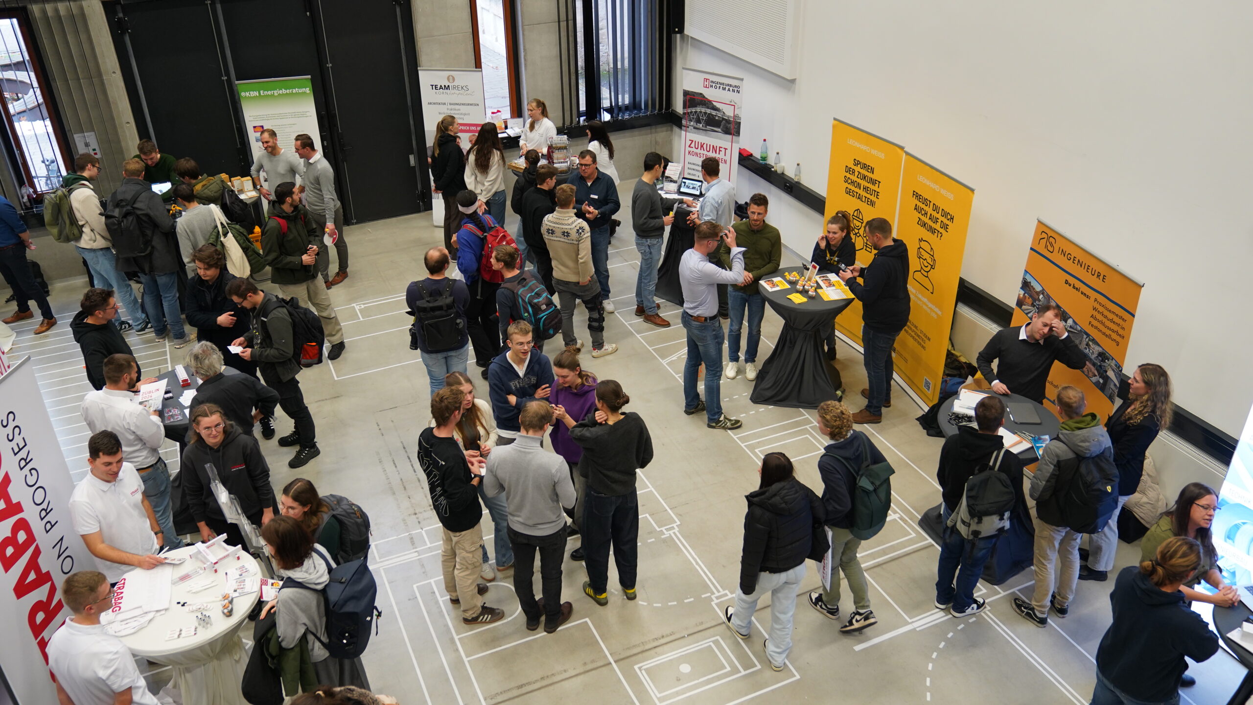 Eine Gruppe von Menschen, darunter auch Nachwuchstalente, versammelt sich an verschiedenen Ständen in einer großen, hellen Halle - wahrscheinlich auf einem Unternehmenstreff oder einer Karrieremesse - und tauscht sich aus. Einige unterhalten sich, während andere die Stände durchstöbern oder Informationsmaterial mitnehmen. at Hochschule Coburg