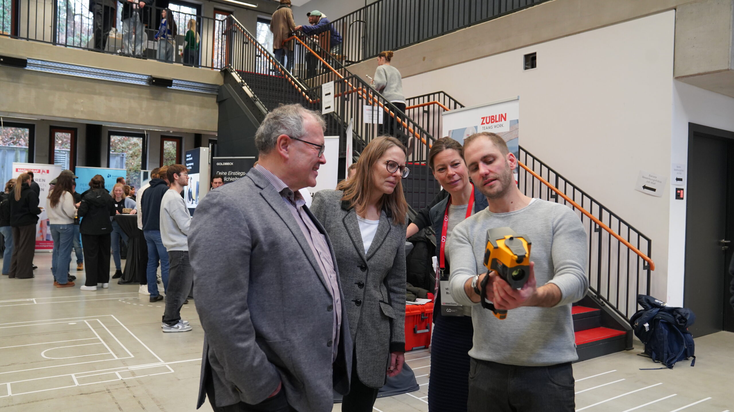 Ein Mann demonstriert drei Nachwuchstalenten in Geschäftskleidung ein gelbes Handheld-Gerät in einem modernen Gebäude mit einem offenen Treppenhaus und anderen Personen, die sich im Hintergrund unter die Leute mischen. at Hochschule Coburg