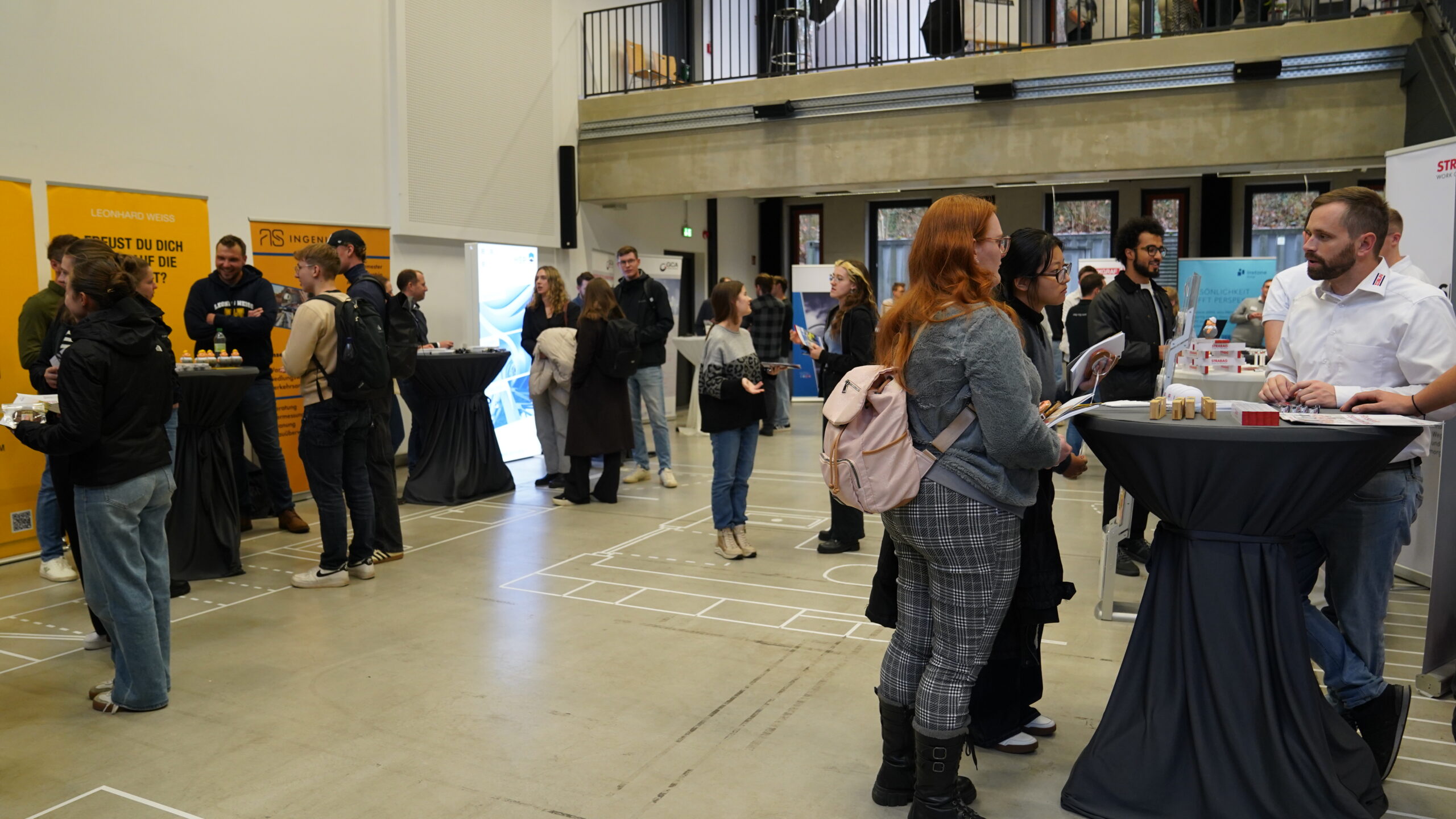 Bei diesem Treff treffen sich Unternehmen und knüpfen Kontakte an hohen Cocktailtischen bei einer Indoor-Veranstaltung. Informationsstände mit Bannern säumen den Raum, während die Teilnehmer, darunter auch Nachwuchstalente, Broschüren in der Hand halten und Rucksäcke tragen. at Hochschule Coburg