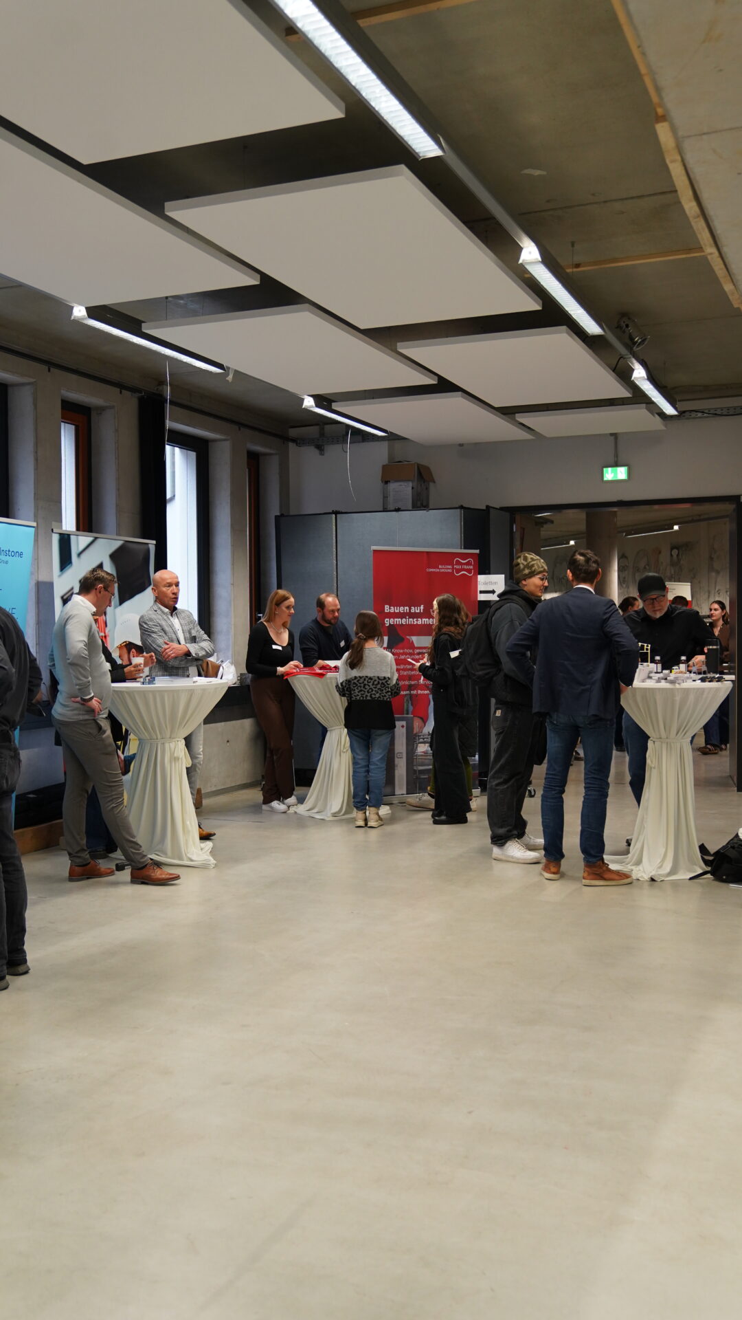 Eine Gruppe von Personen steht um Stehtische herum und unterhält sich bei einer Indoor-Veranstaltung von Unternehmen. Einige lesen Broschüren oder benutzen Laptops; im Hintergrund sind Banner und Plakate zu sehen, die eine moderne, professionelle Atmosphäre schaffen, die ideal für den Aufbau von Kontakten ist. at Hochschule Coburg